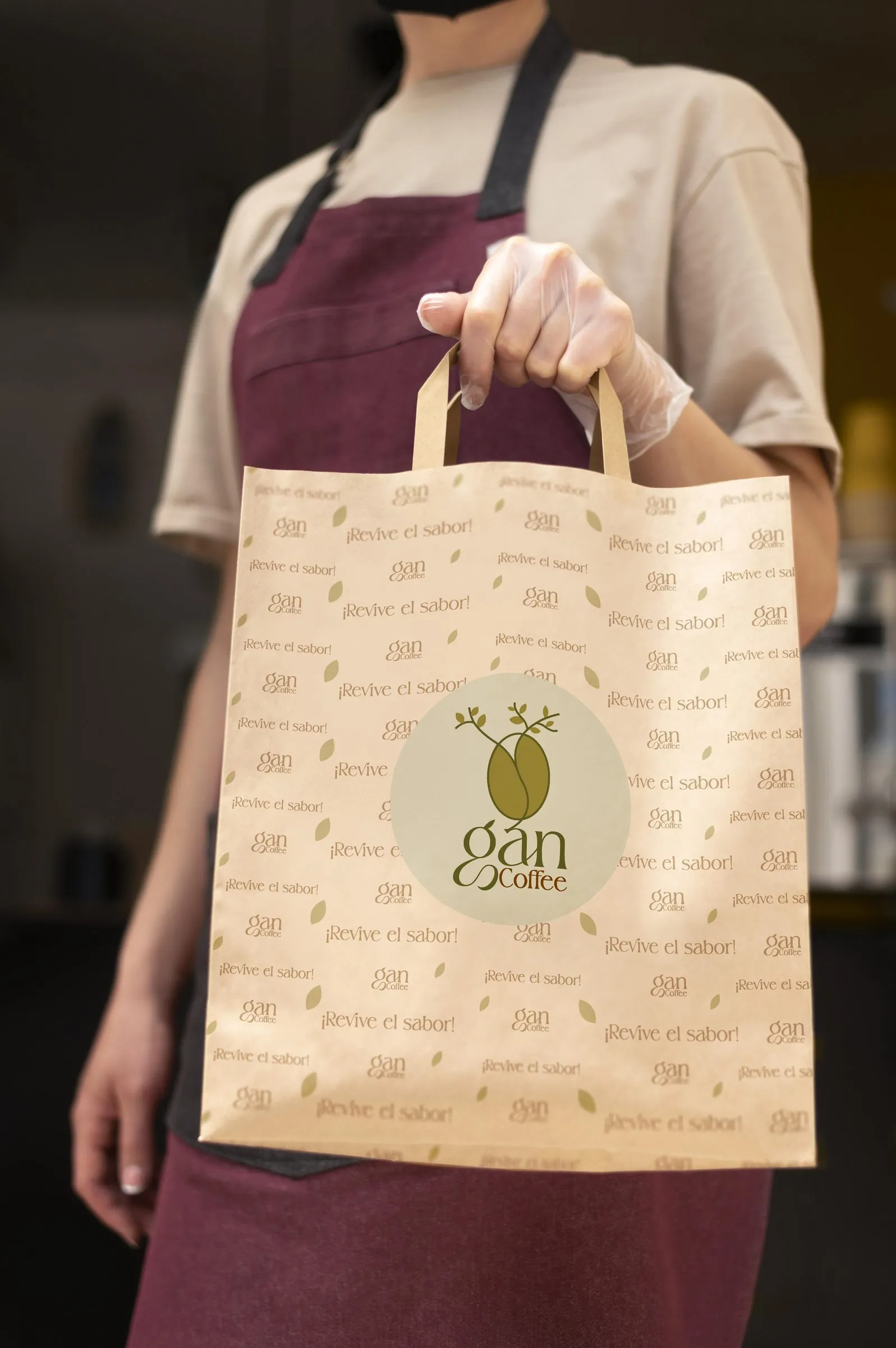 Persona con delantal vino y guante desechable sosteniendo una bolsa de papel kraft con el logo de Gan Coffee y el texto ¡Revive el sabor! repetido.