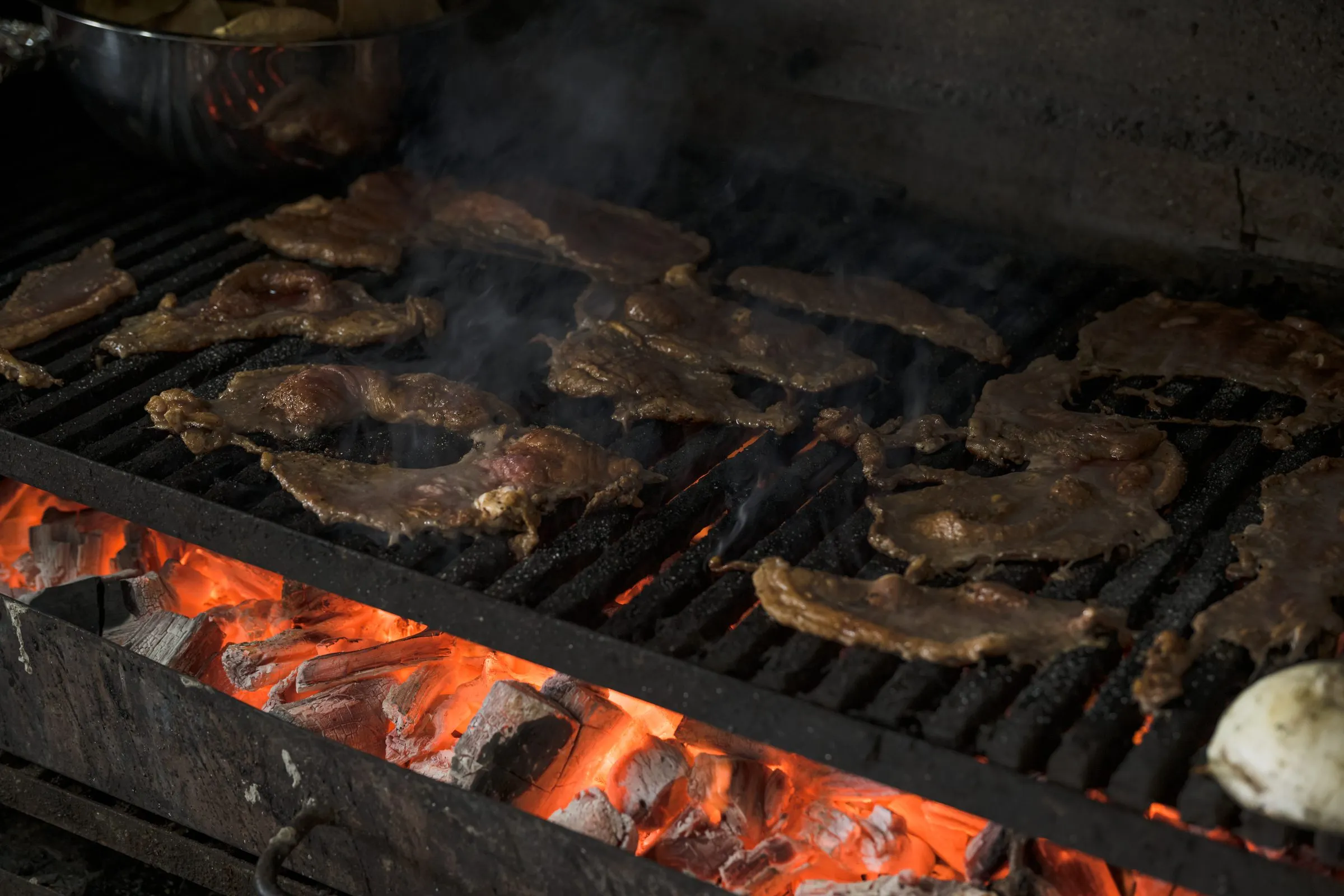 Carne sobre parrilla de brasas encendidas, humo subiendo, el proceso de cocción visible de una taquería real