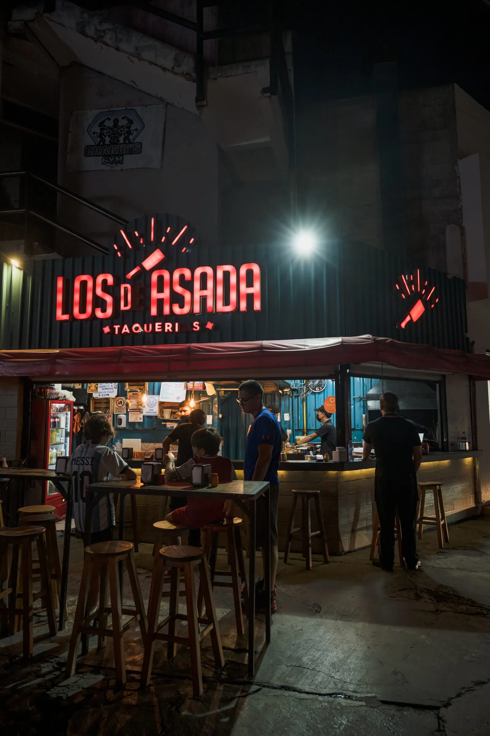 Fachada nocturna de la taquería con letrero de neón encendido, mesas altas y clientes en la barra, el local tal como es