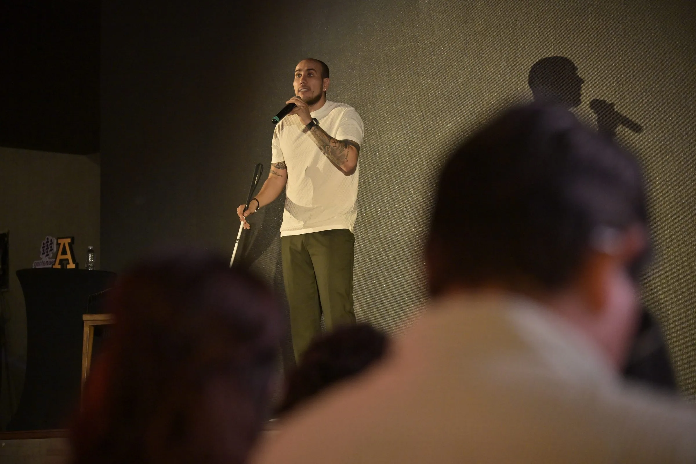 Backlit speaker mid-gesture on stage with the audience visible in silhouette