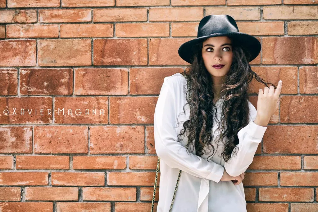 Mujer con sombrero y camisa blanca recargada en un muro de ladrillo