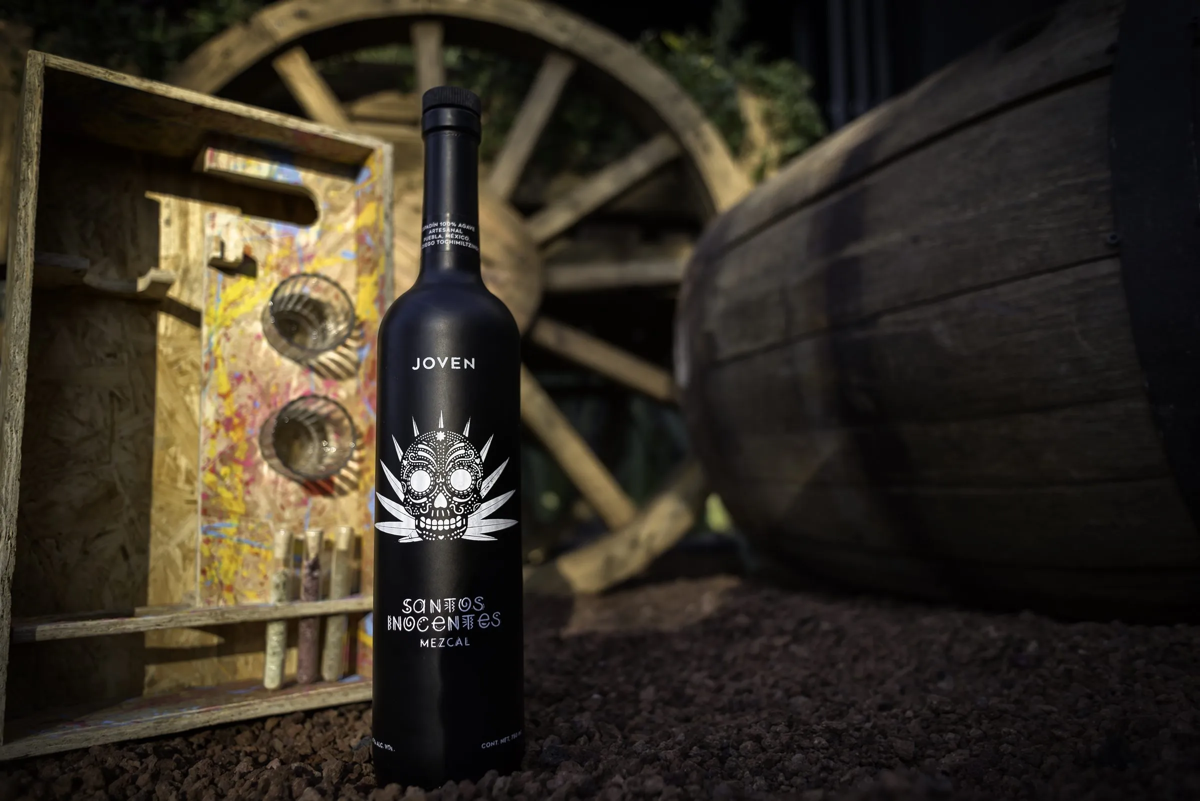 Botella de mezcal Santos Inocentes Joven, negra mate con una calavera decorada entre hojas de agave, de pie sobre tierra oscura frente a una rueda de carreta de madera