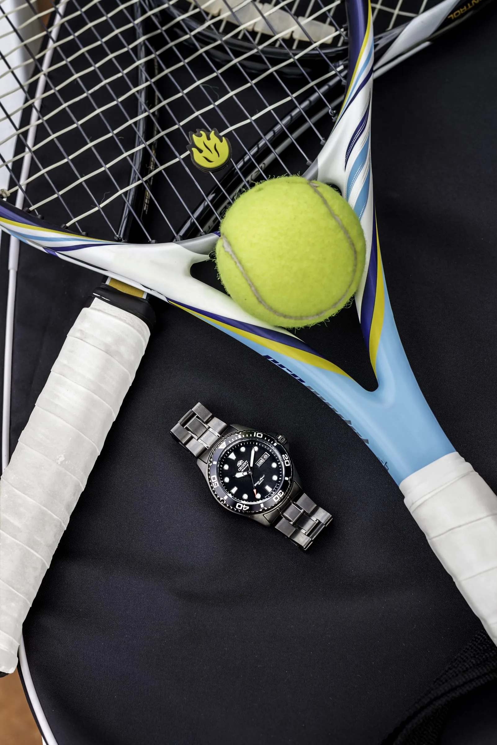Black-dial Orient watch placed beside a tennis racket and ball on a dark sports bag, shot from above.