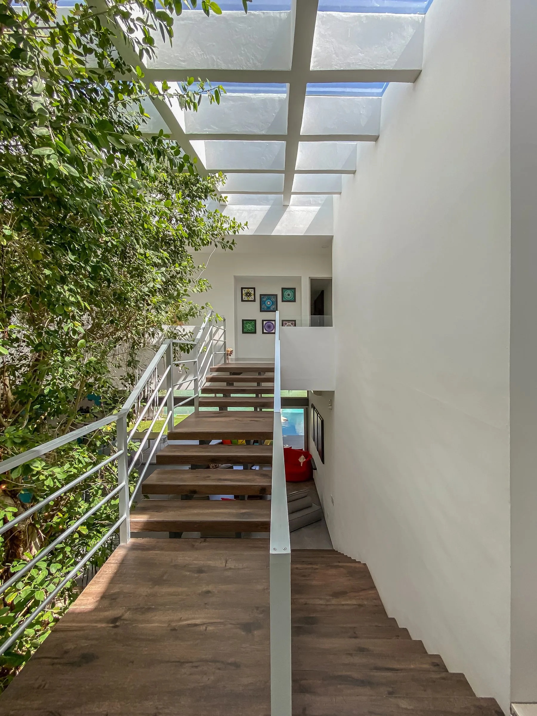 Escalera de madera exterior junto a un árbol, bajo un tragaluz con estructura blanca, con barandal metálico y platos decorativos enmarcados al fondo, y una alberca visible más abajo.