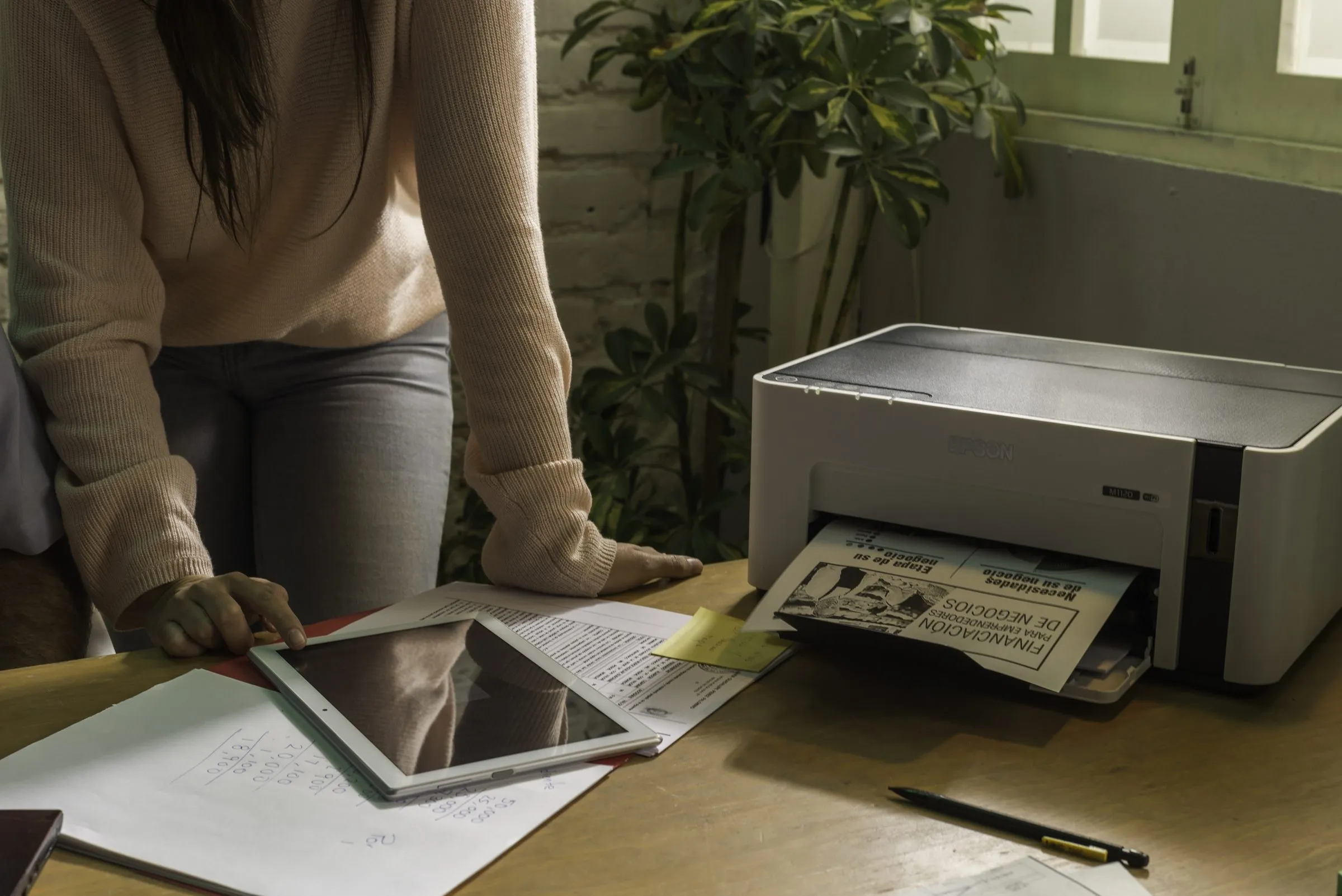 Persona junto a una impresora que expulsa una hoja impresa, con una tableta y documentos sobre el escritorio y una planta al fondo.