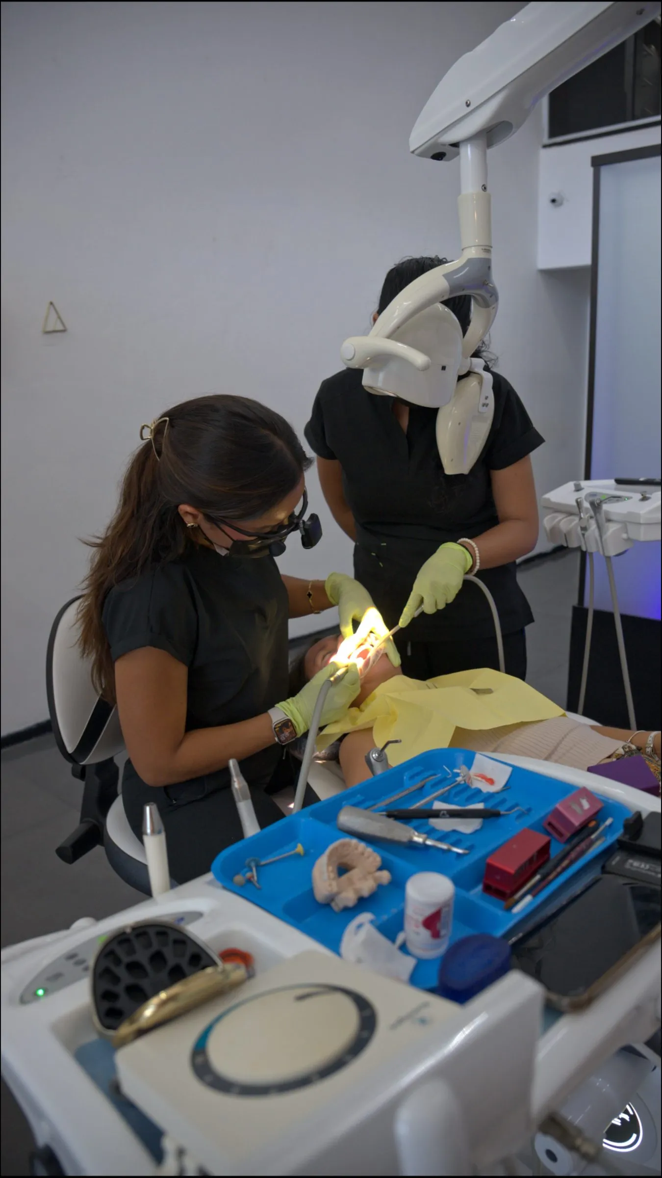 Dentista y asistente con guantes y lentes de protección atienden a un paciente en el sillón dental, con instrumental y una lámpara cenital encendida.