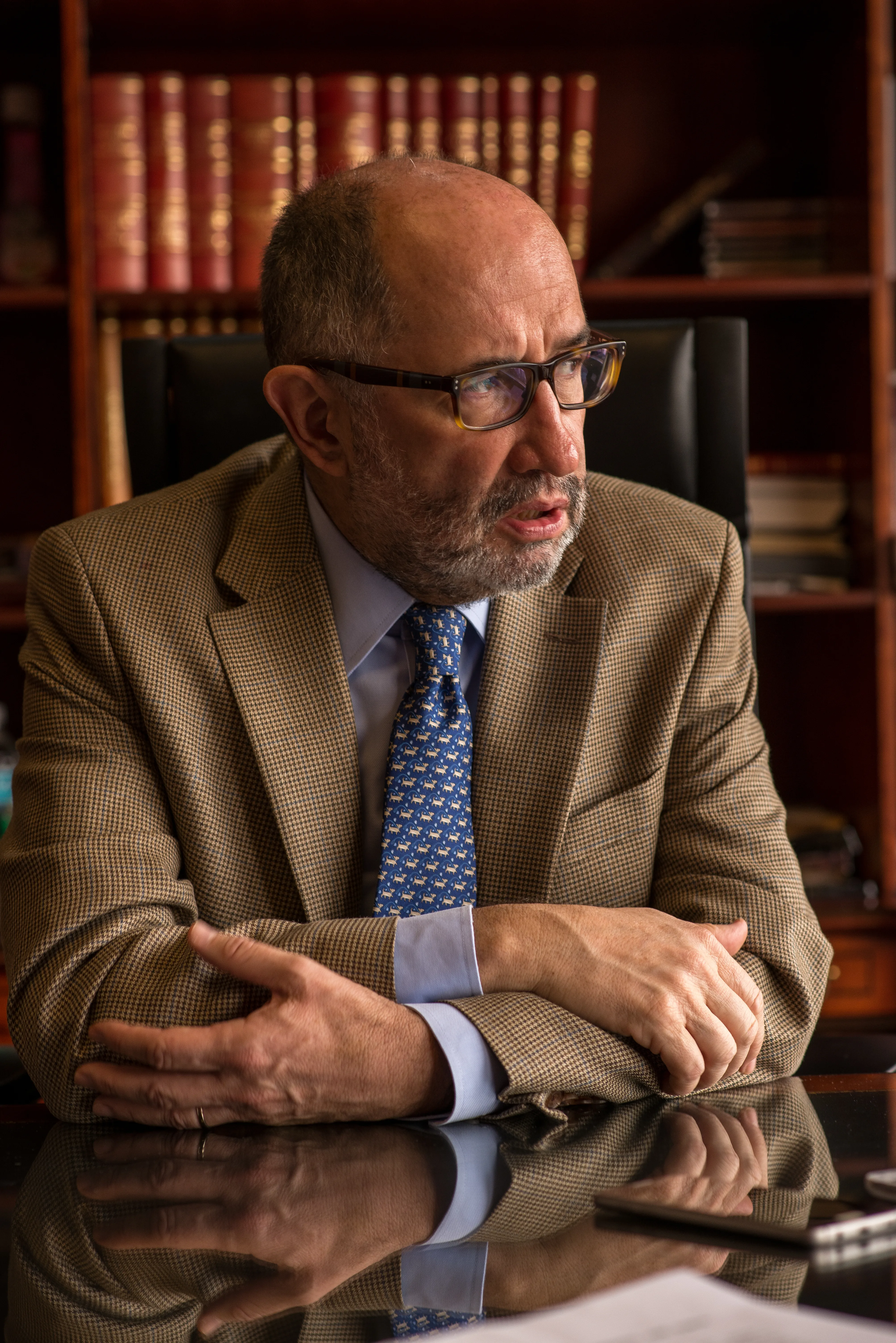 Retrato de un jurista de saco de espiga sentado con los brazos cruzados sobre su escritorio, con libros de derecho al fondo
