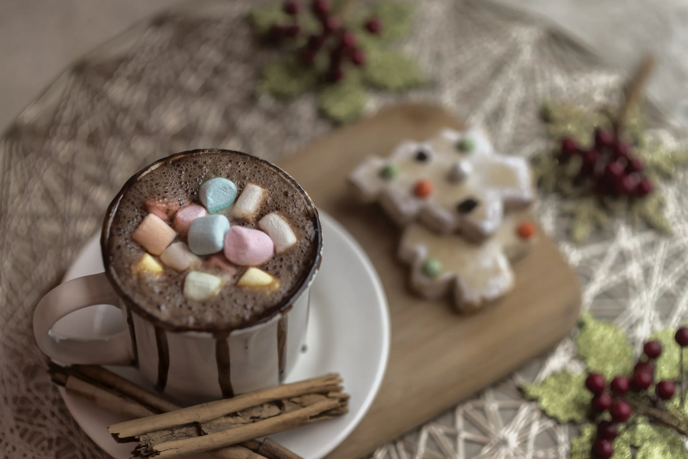 Taza de chocolate caliente con malvaviscos de colores y chocolate escurriendo por las paredes, servida sobre un plato con rajas de canela y una galleta decorada en forma de estrella de fondo.