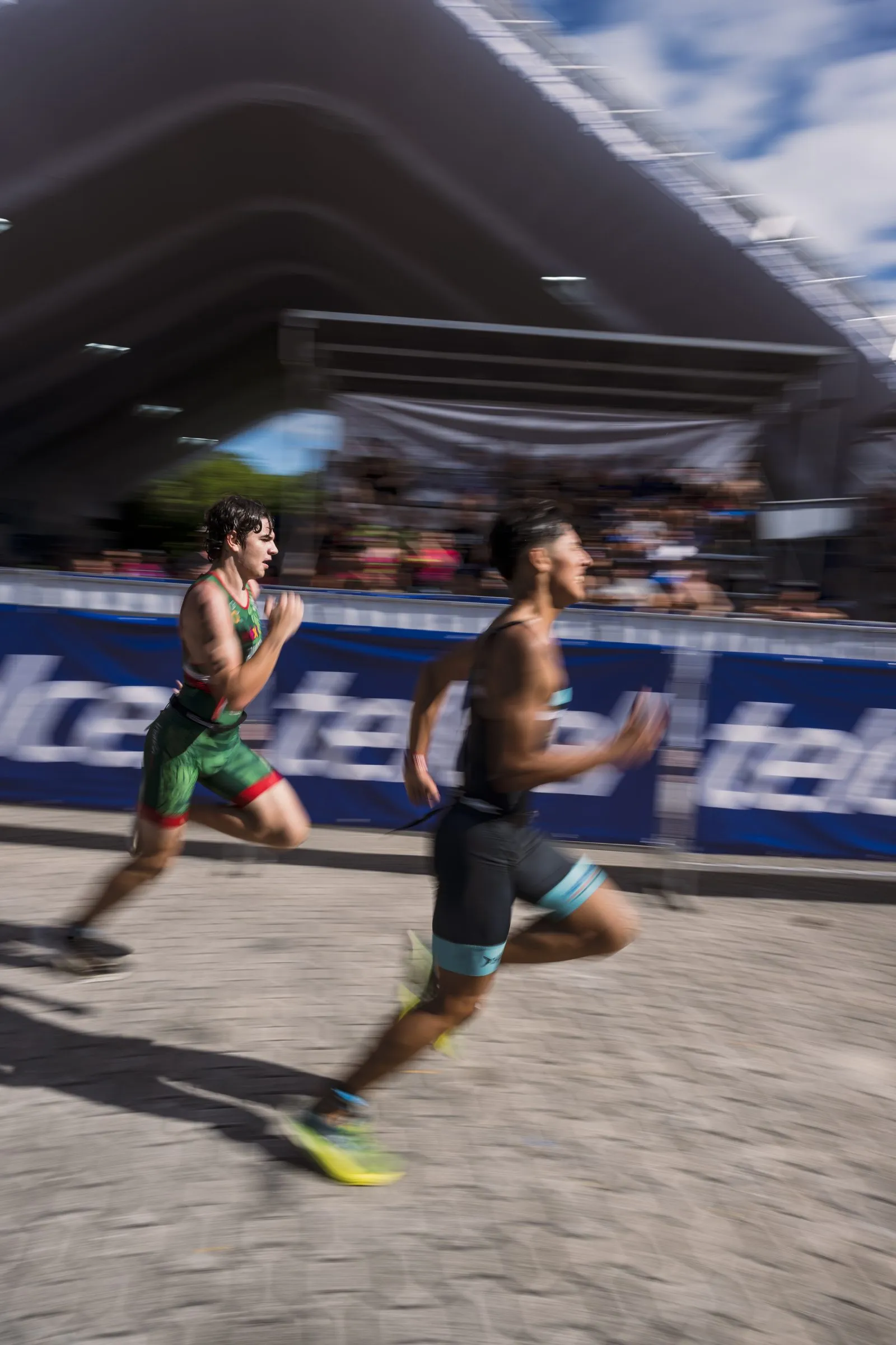 Two triathletes run side by side mid-race, captured with motion blur, in front of a Telcel-branded barrier with spectators in the stands behind them.