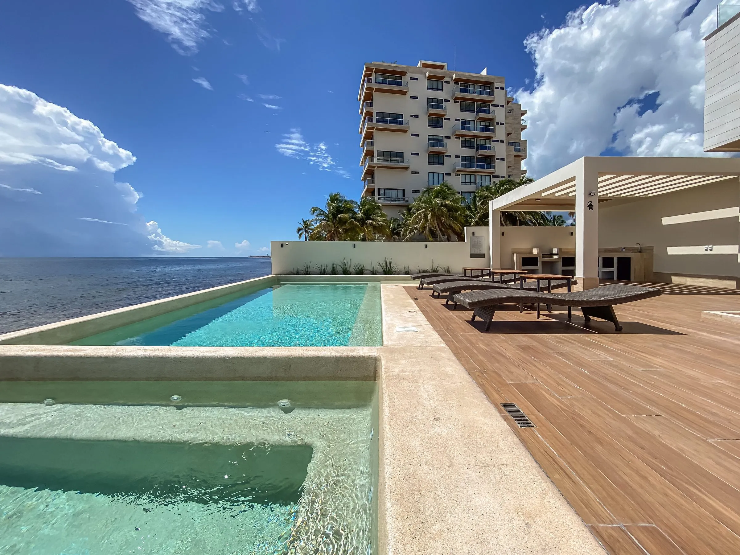 Terraza de alberca infinita junto al mar, con camastros de madera, un jacuzzi en primer plano, palapa con asador y un edificio residencial entre palmeras al fondo.