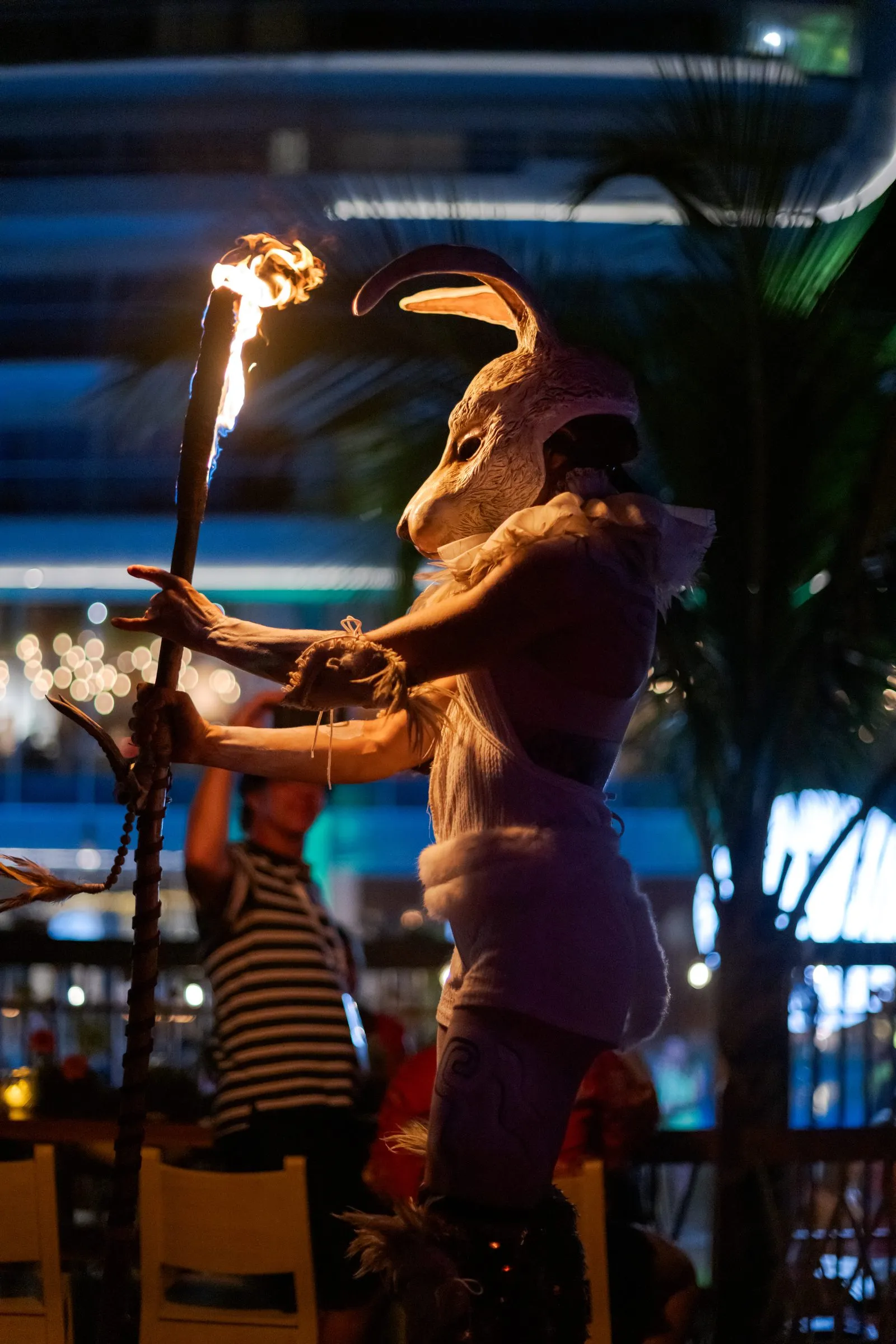 Artista con disfraz y máscara de liebre sostiene un bastón encendido durante una presentación nocturna, con luces de neón y palmeras al fondo.