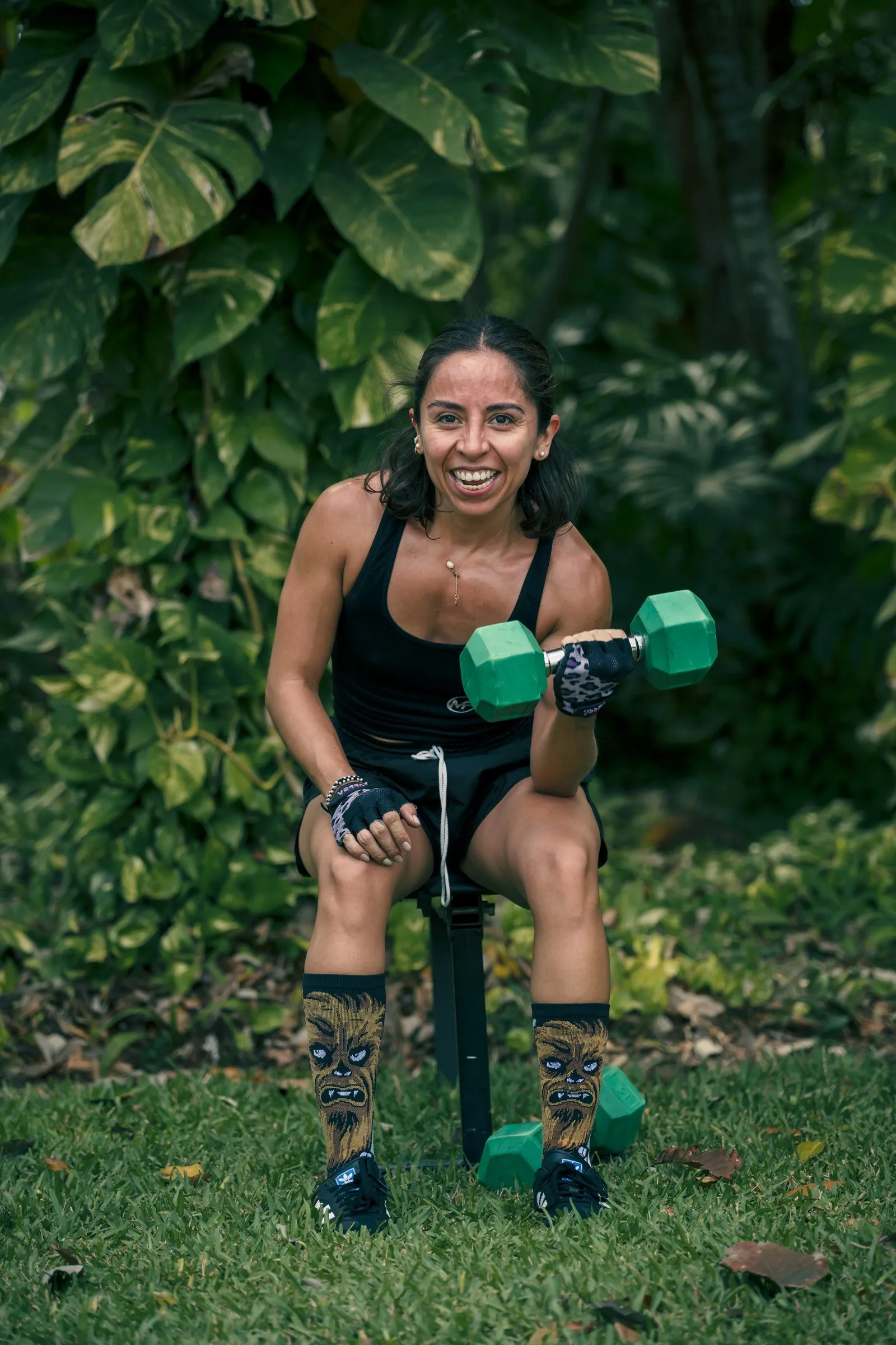 Mujer sentada en una banca al aire libre rodeada de follaje verde, haciendo un curl de bíceps con una mancuerna verde y otra mancuerna apoyada en el pasto.