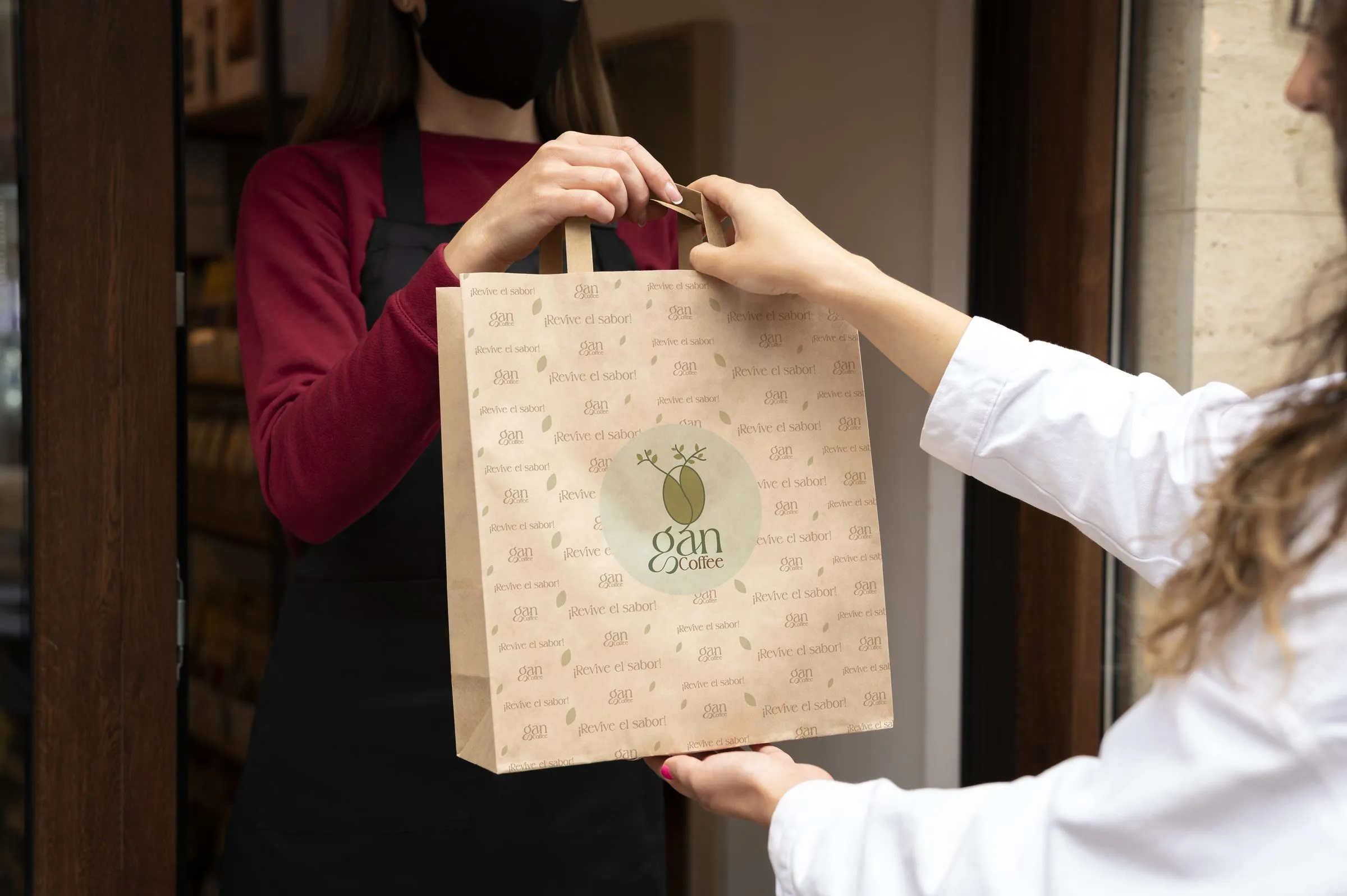 Dos personas en una puerta intercambiando la bolsa de papel kraft de Gan Coffee, una con cubrebocas negro y delantal, la otra tomando las asas con mangas blancas.