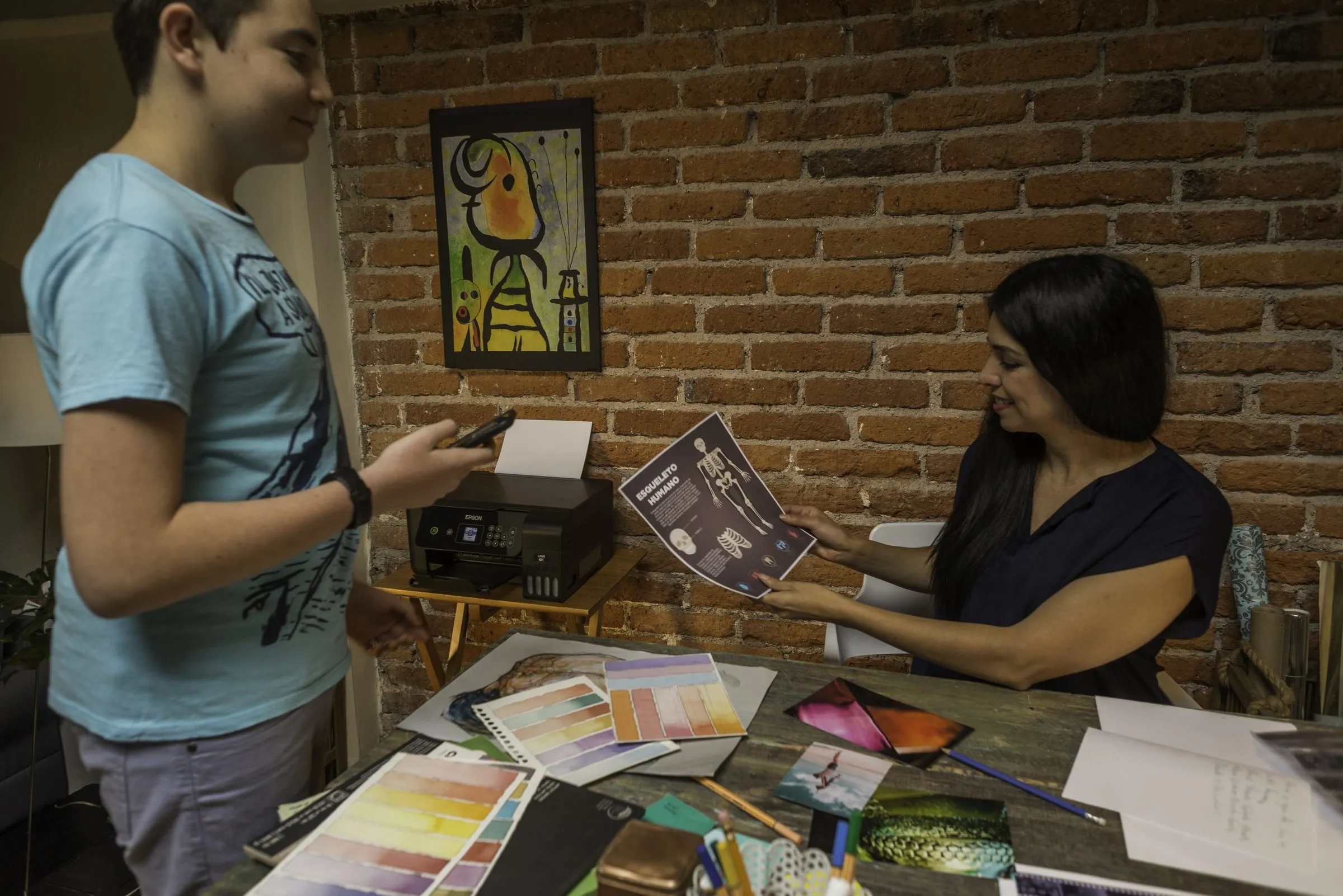 Un adolescente y una mujer revisan una hoja impresa sobre el esqueleto humano junto a una impresora, con muestras de acuarela sobre la mesa.