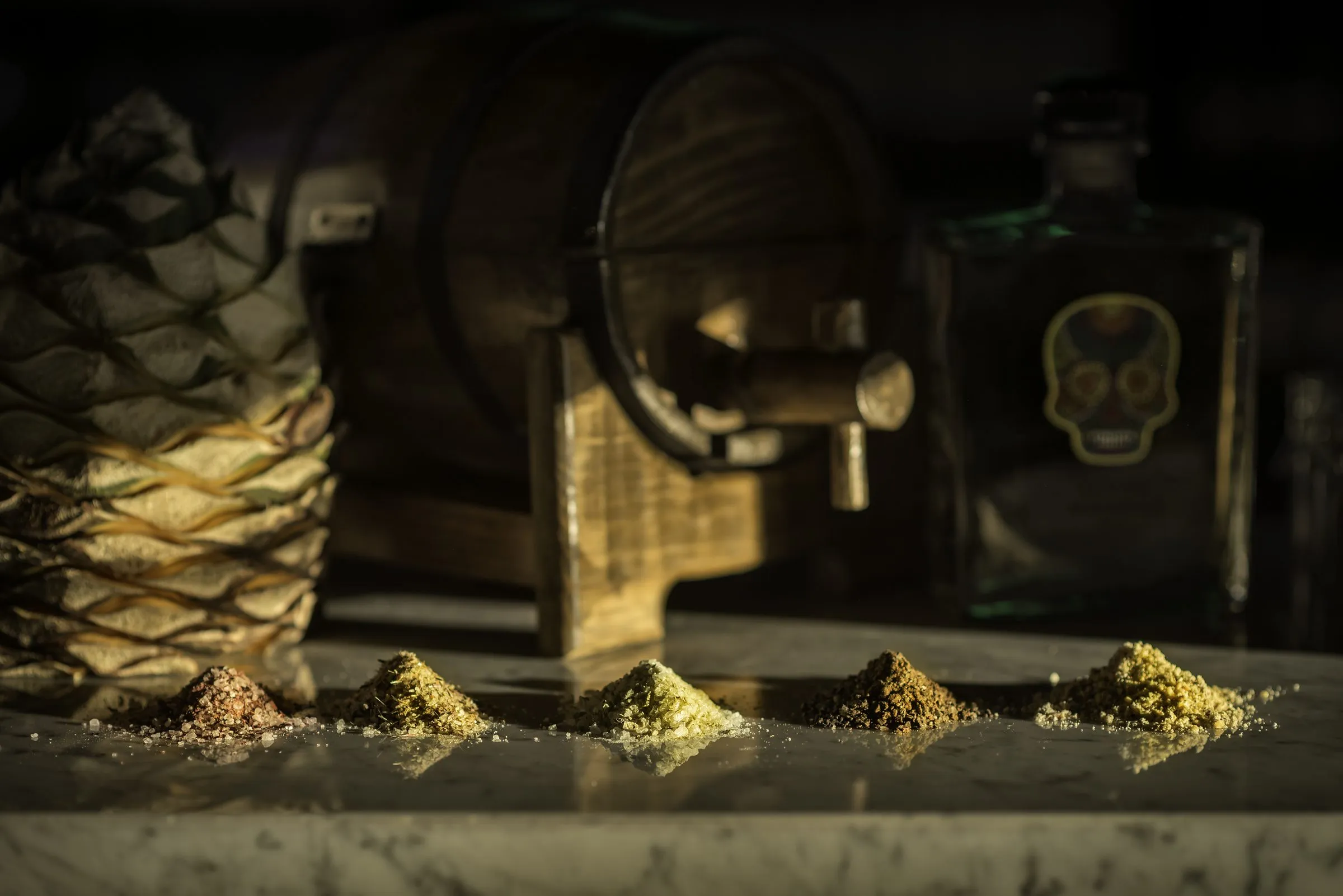 Cinco montículos de sal de colores distintos alineados sobre mármol, con una piña de agave asada y una botella con etiqueta de calavera desenfocadas al fondo