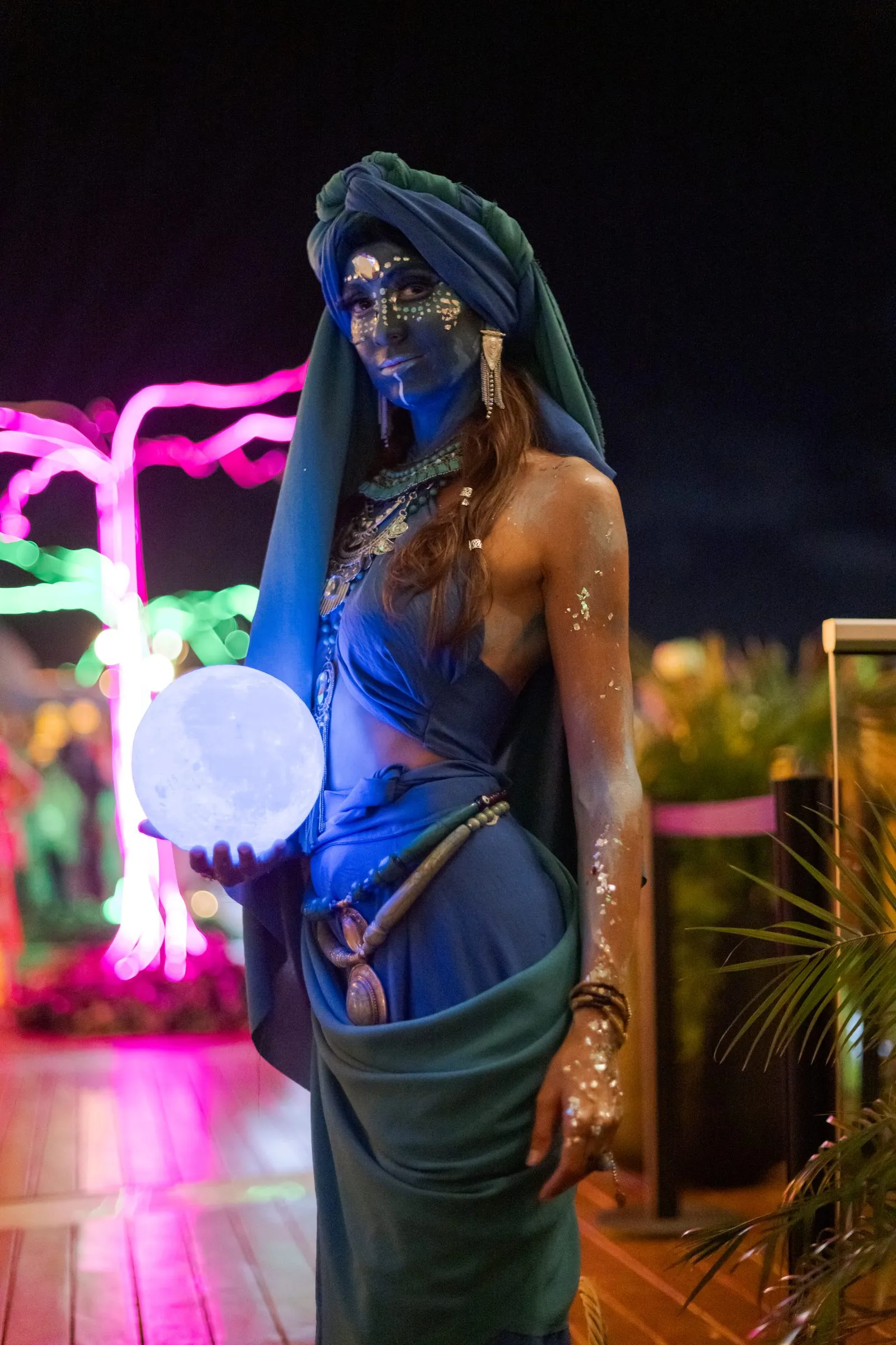 Artista con maquillaje corporal azul, tocado y joyería elaborados, sostiene una esfera luminosa entre instalaciones de luces de neón.