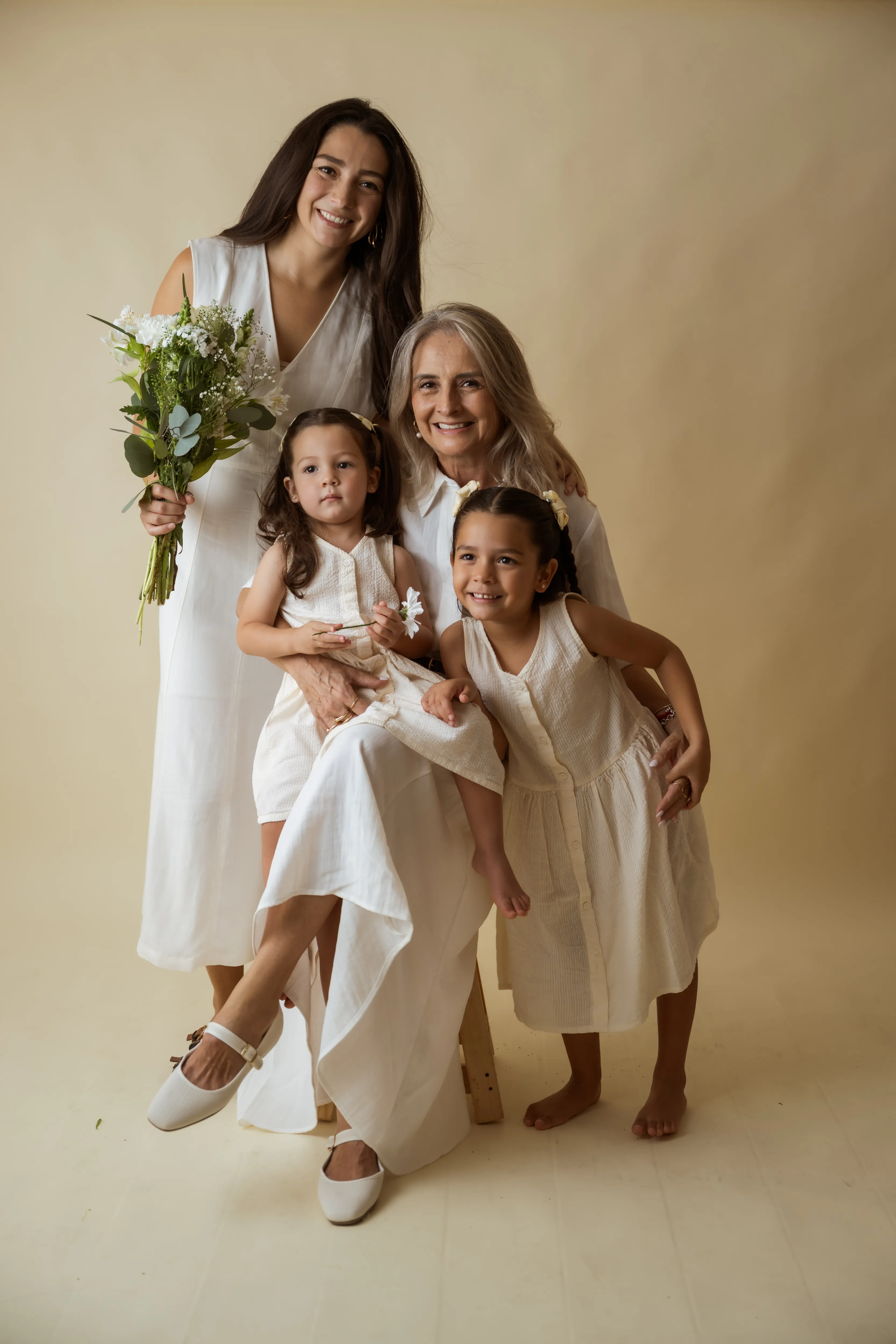 Tres generaciones vestidas de blanco — abuela, mamá y niñas — con un ramo de flores en set de estudio.