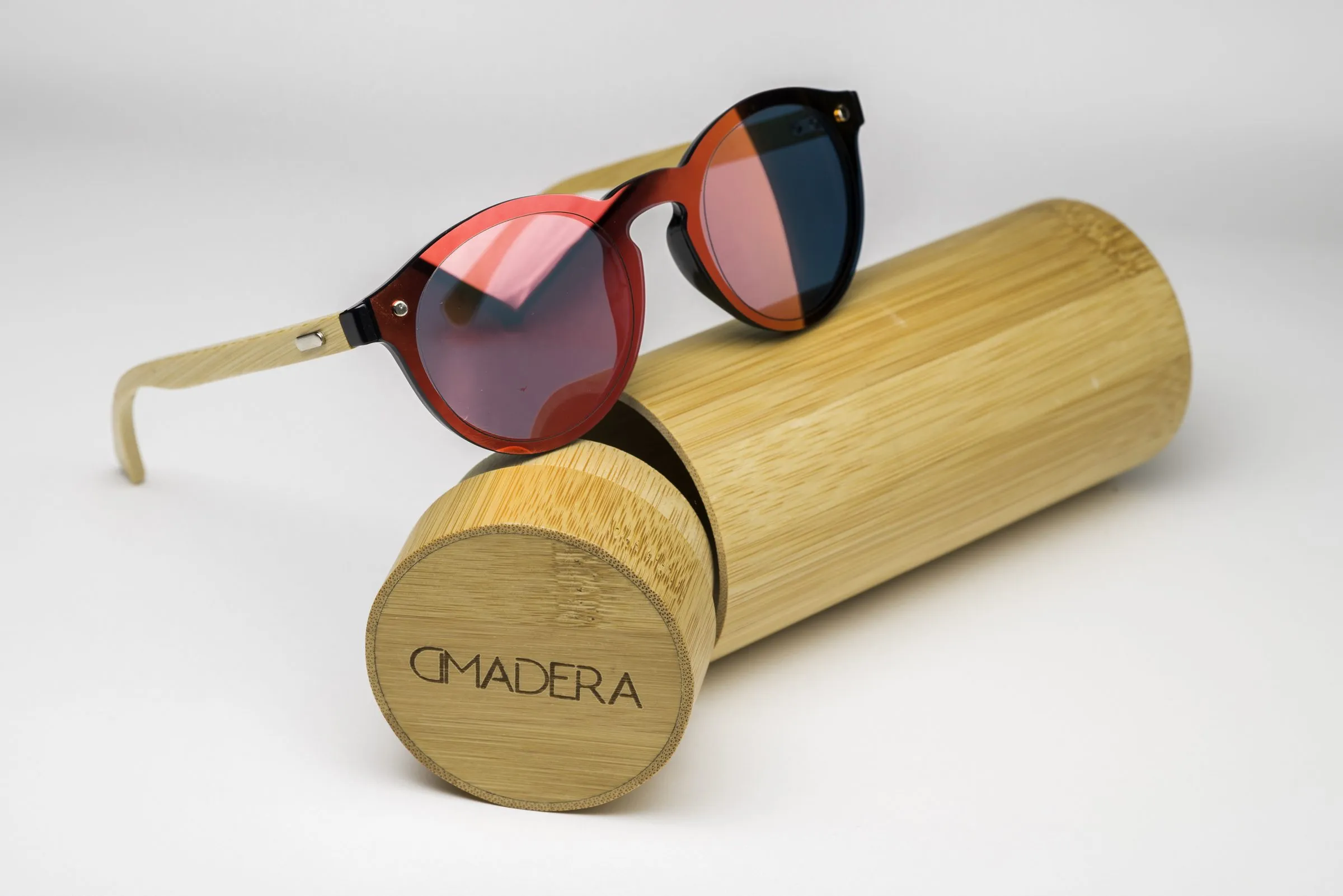 Round sunglasses with a red tortoiseshell-toned front and lenses mirrored from pink to blue, wooden temple arms, resting on a cylindrical wooden case with the brand logo engraved on the end.