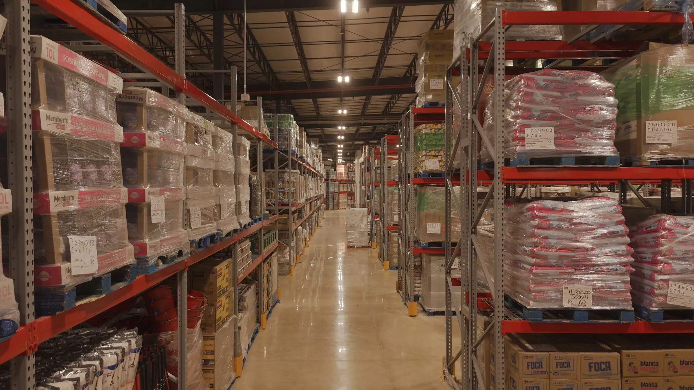 Warehouse aisle lined with racks holding shrink wrapped pallets of cleaning products and toilet paper above a polished concrete floor