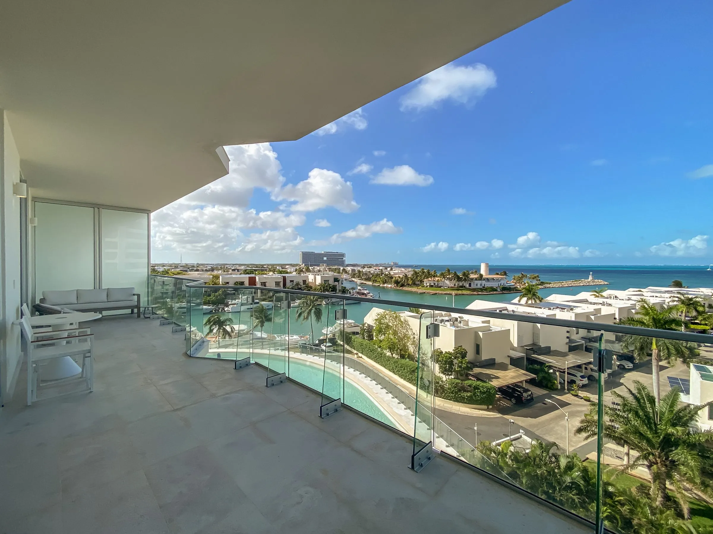 Terraza con barandal de vidrio y muebles de exterior, con vista a una marina con yates, palmeras y edificios blancos frente al mar bajo un cielo azul con nubes.