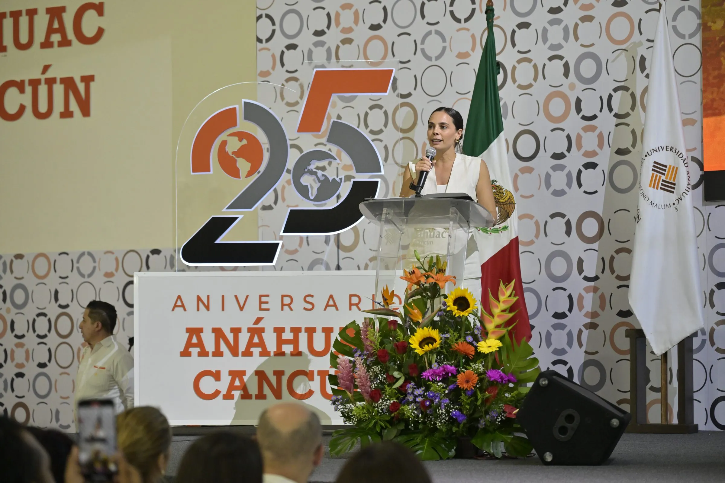Mujer con vestido blanco habla desde un podio junto al logotipo del 25 aniversario, con arreglo floral en primer plano y asistentes desenfocados en primer término.