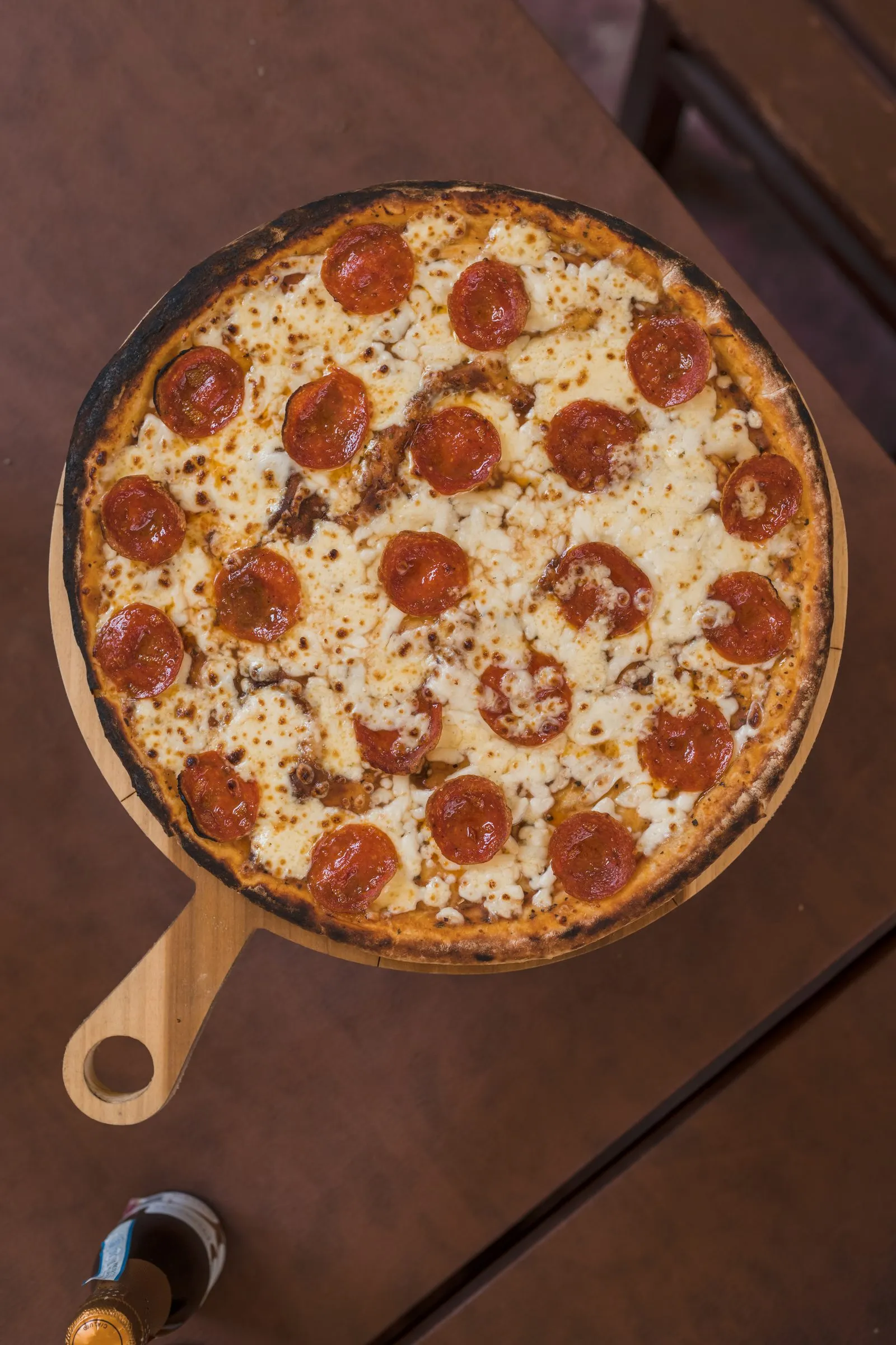 Pizza de pepperoni con queso derretido y orilla ligeramente quemada, servida entera sobre una tabla de madera redonda con mango.