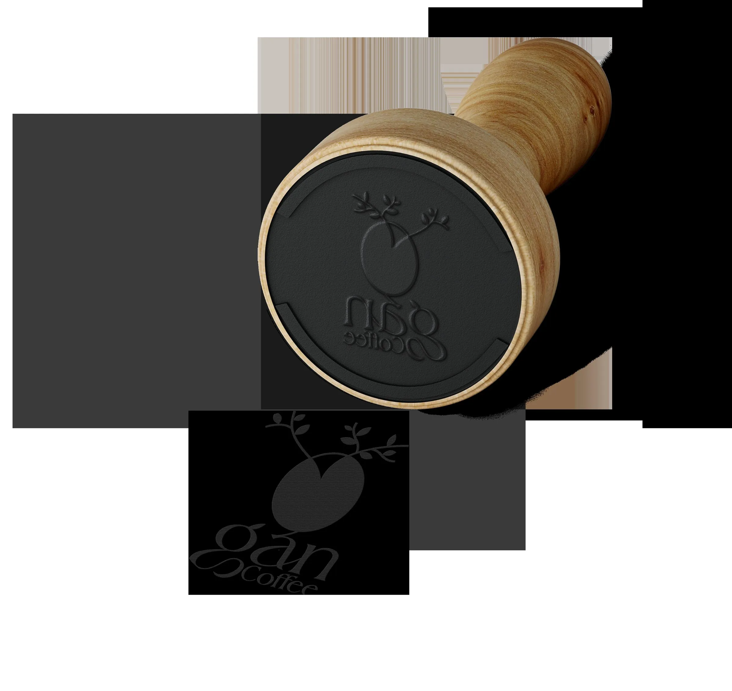 Pisón de café de madera con la cara plana grabada con el logo de hoja y grano de Gan Coffee, en ángulo sobre fondo oscuro, con el logotipo repetido abajo en un recuadro negro.