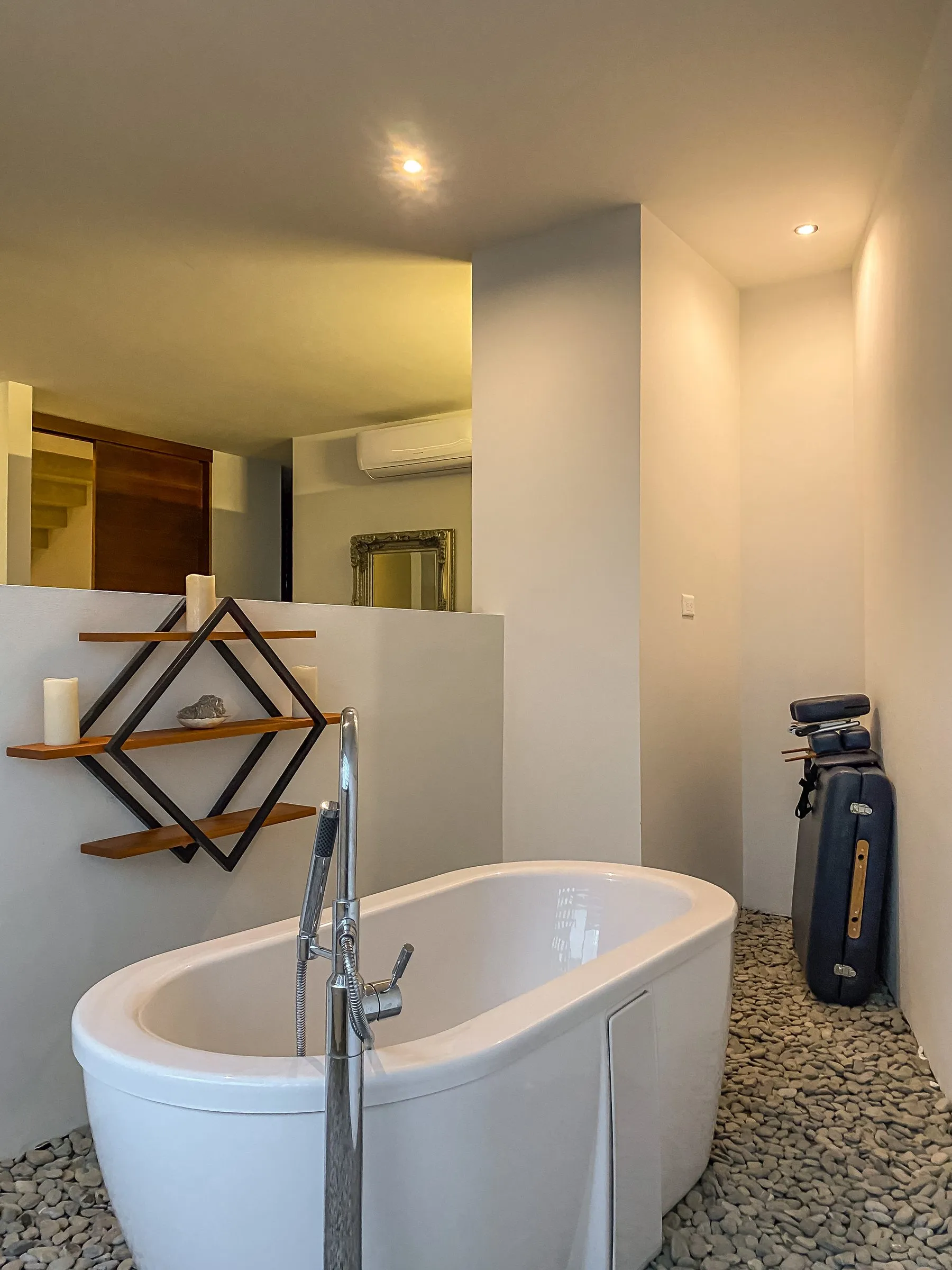 Baño con tina ovalada independiente sobre piso de piedra río, grifo cromado de piso, un estante en forma de rombo con velas, y vista a una escalera y un espejo con marco ornamentado.