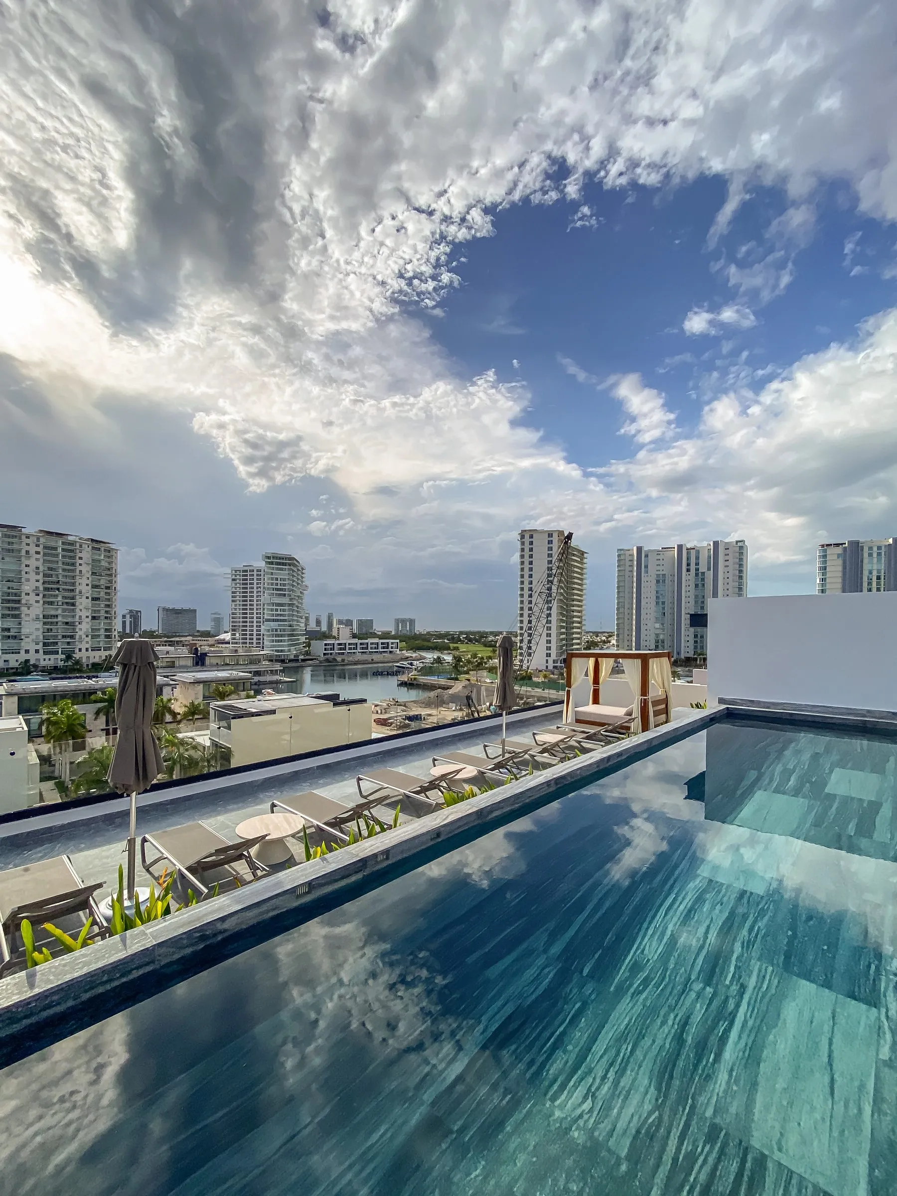Alberca infinita en una azotea con camastros, sombrillas y una cama balinesa, con vista panorámica de la marina y los edificios de la ciudad reflejados en el agua.