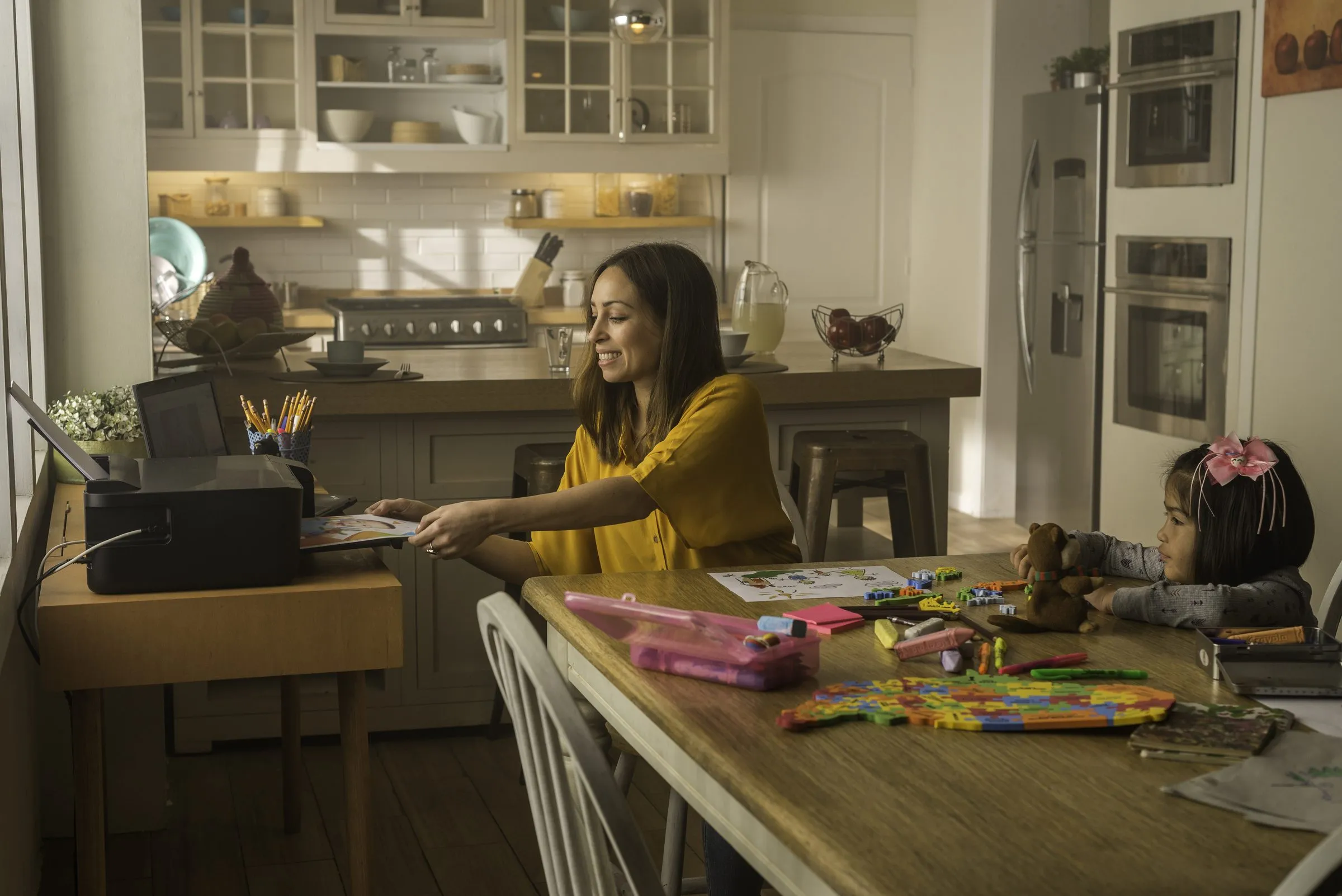 En una cocina luminosa, una mujer entrega una hoja impresa a una niña sentada a la mesa con crayones y un peluche.