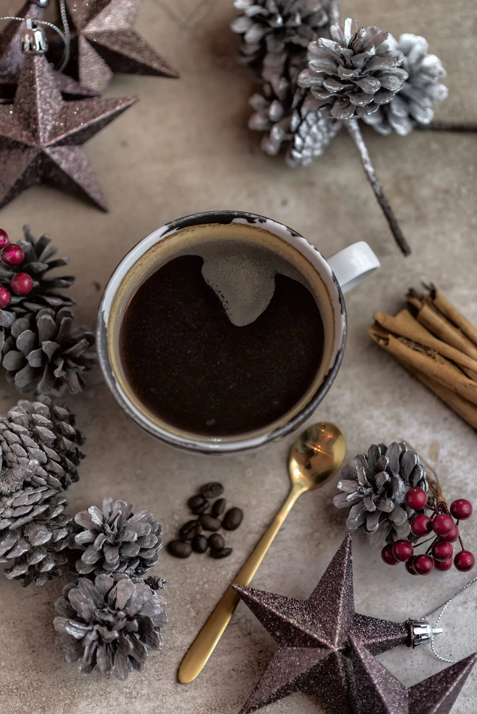 Café negro en una taza con diseño blanco y negro, fotografiado desde arriba junto a granos de café, canela y piñas plateadas sobre una superficie de concreto.