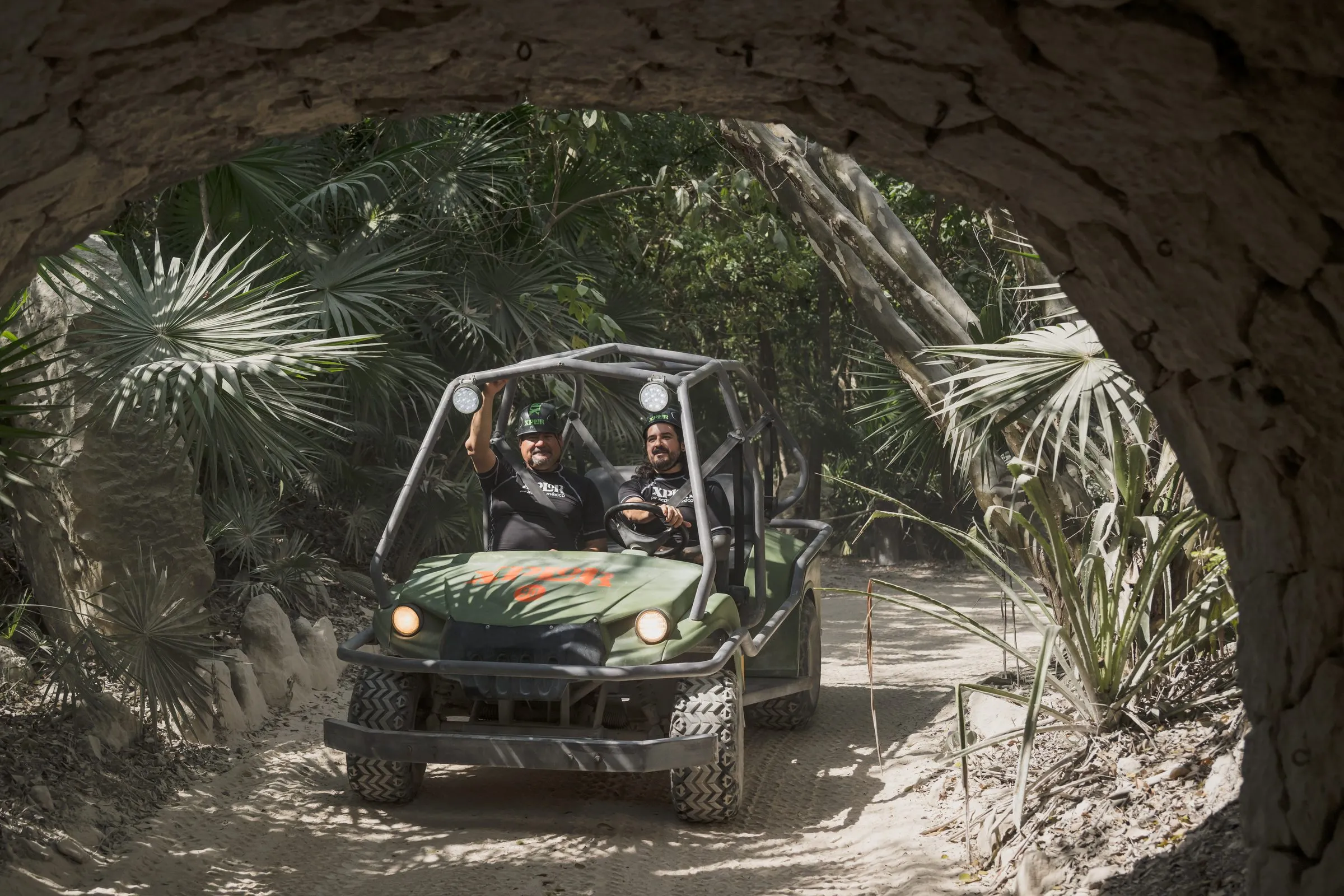 Dos hombres con casco conducen un vehículo todoterreno con el logo de Xplor por un túnel de piedra rodeado de vegetación selvática.