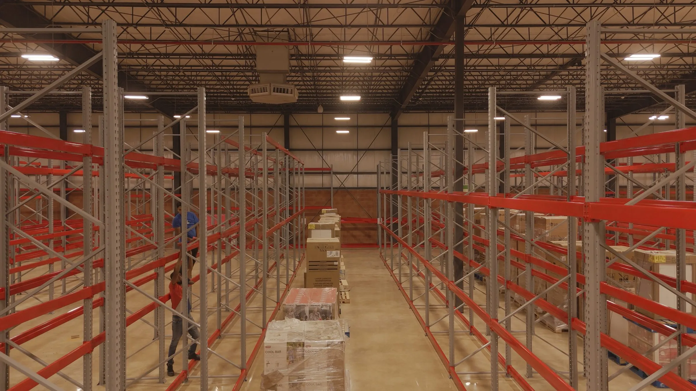 Aisle of industrial pallet racking with red beams, boxed appliances stacked on pallets in the center walkway, and two workers in the aisle to the left