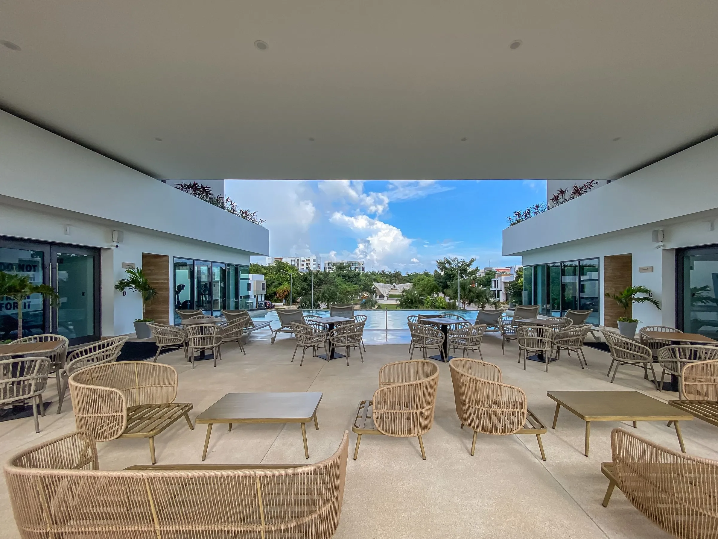 Covered terrace with rattan armchairs and tables facing a pool, greenery and buildings visible beyond under a cloudy sky.