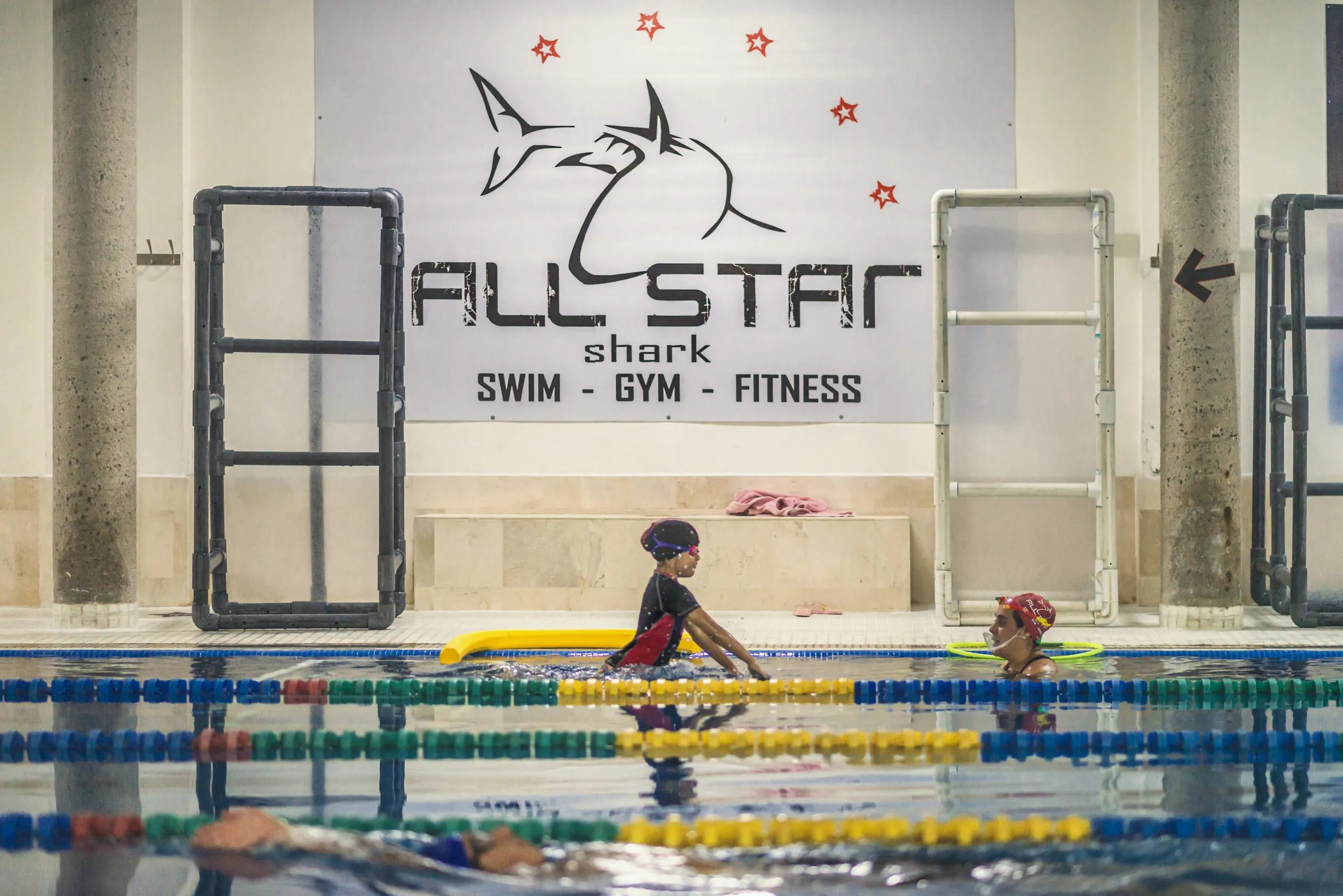Dos alumnos infantiles durante una clase de natación en la alberca del gimnasio, bajo el letrero de la marca All Star