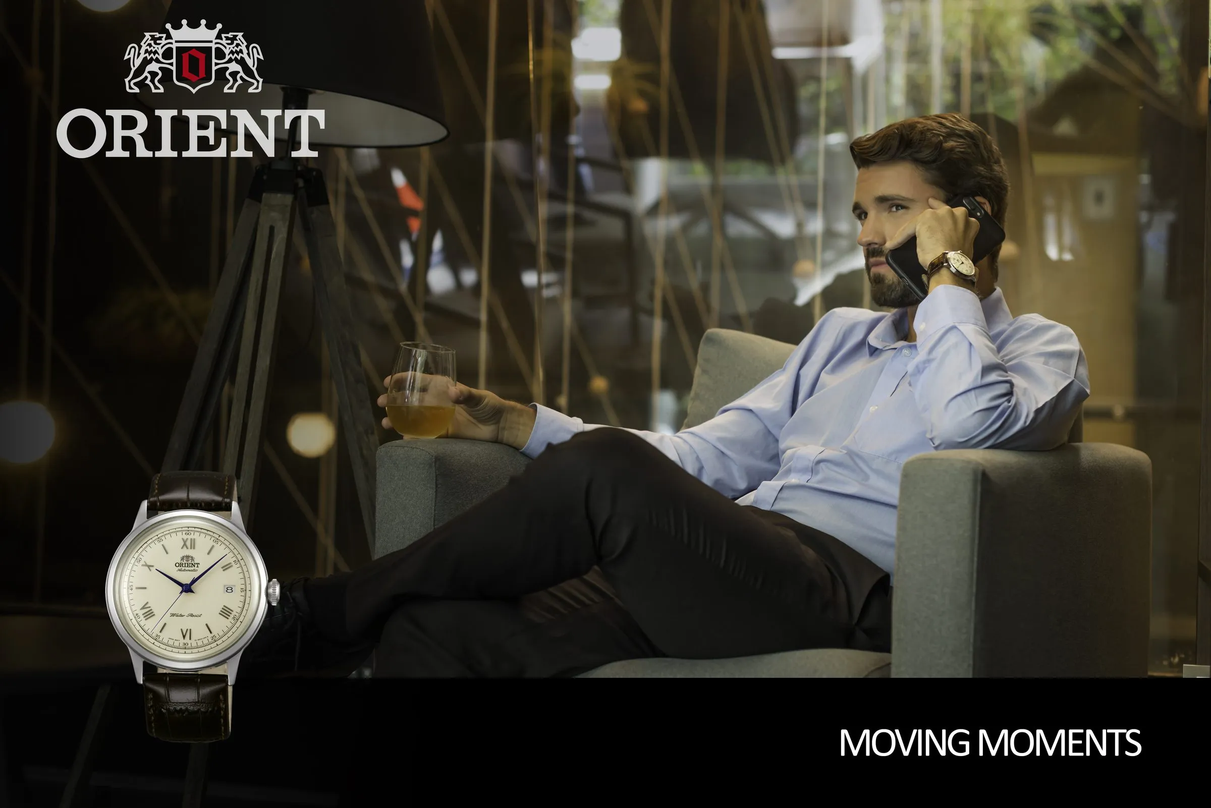 Cream-dial Orient watch in close-up, with a man reclining in an armchair holding a drink and talking on the phone in the background, and the brand's logo visible in the composition.