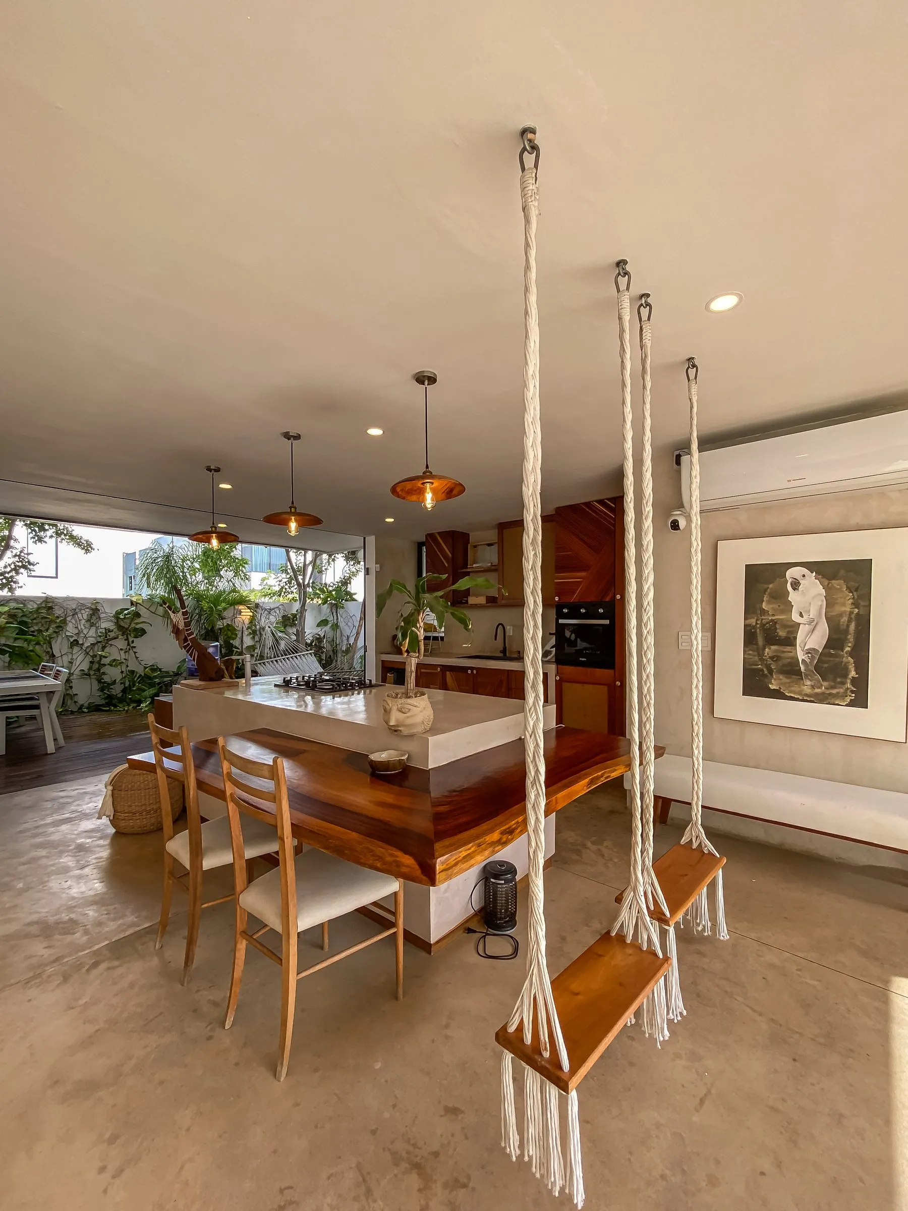 Cocina y comedor abiertos con mesa de madera de borde vivo, cuatro sillas tapizadas, tres lámparas colgantes de cobre sobre una isla de concreto, y columpios de macramé colgados del techo en primer plano.