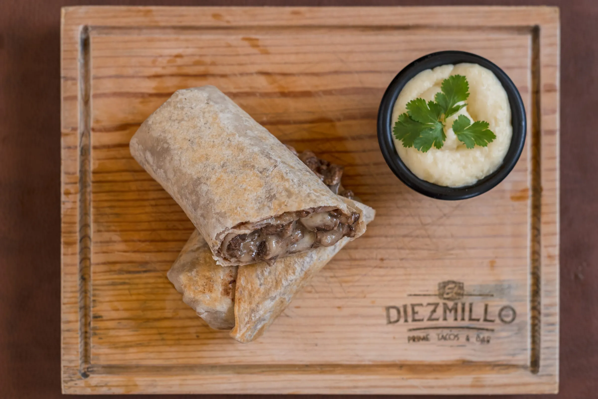 Burrito de carne y queso cortado por la mitad sobre una tabla con el logotipo de Diezmillo, acompañado de un tazón de salsa blanca decorado con cilantro.