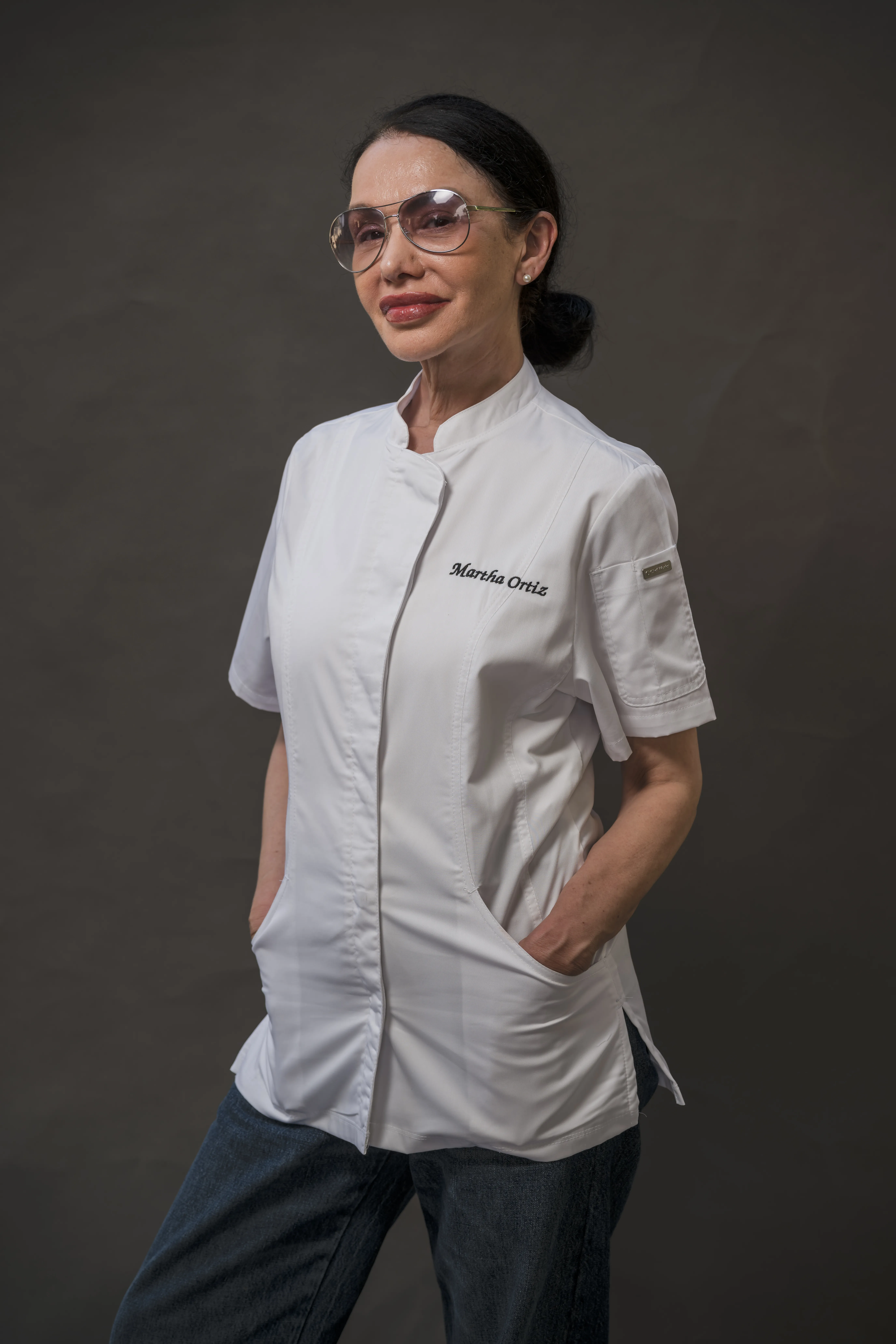 Retrato de estudio de una chef con lentes oscuros y chaqueta blanca bordada con el nombre 'Martha Ortiz', sobre fondo gris.