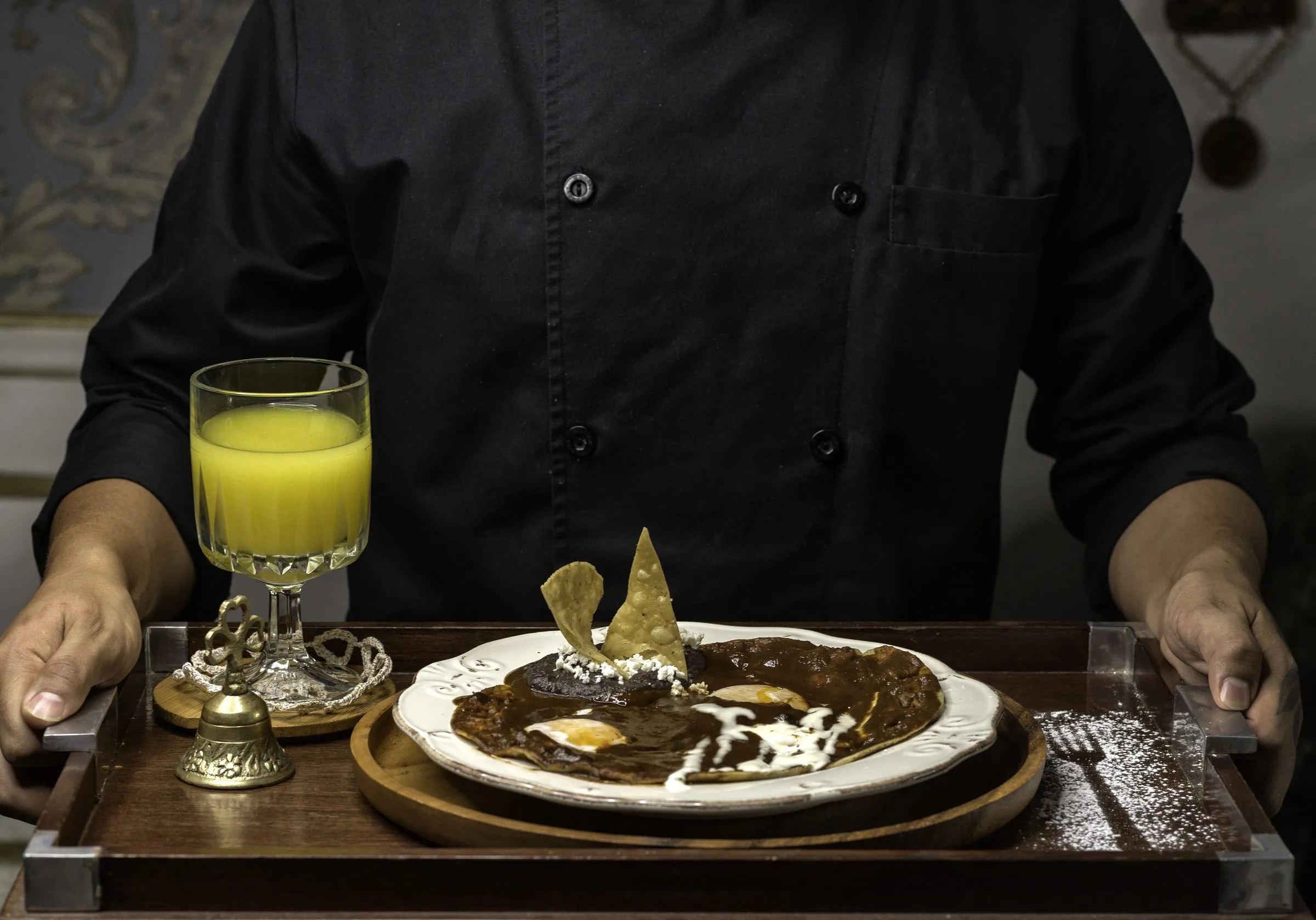 Persona con uniforme negro presentando una charola con un plato de tortillas bañadas en salsa roja con huevo, crema y totopos, además de un vaso de jugo de naranja y una campana decorativa.