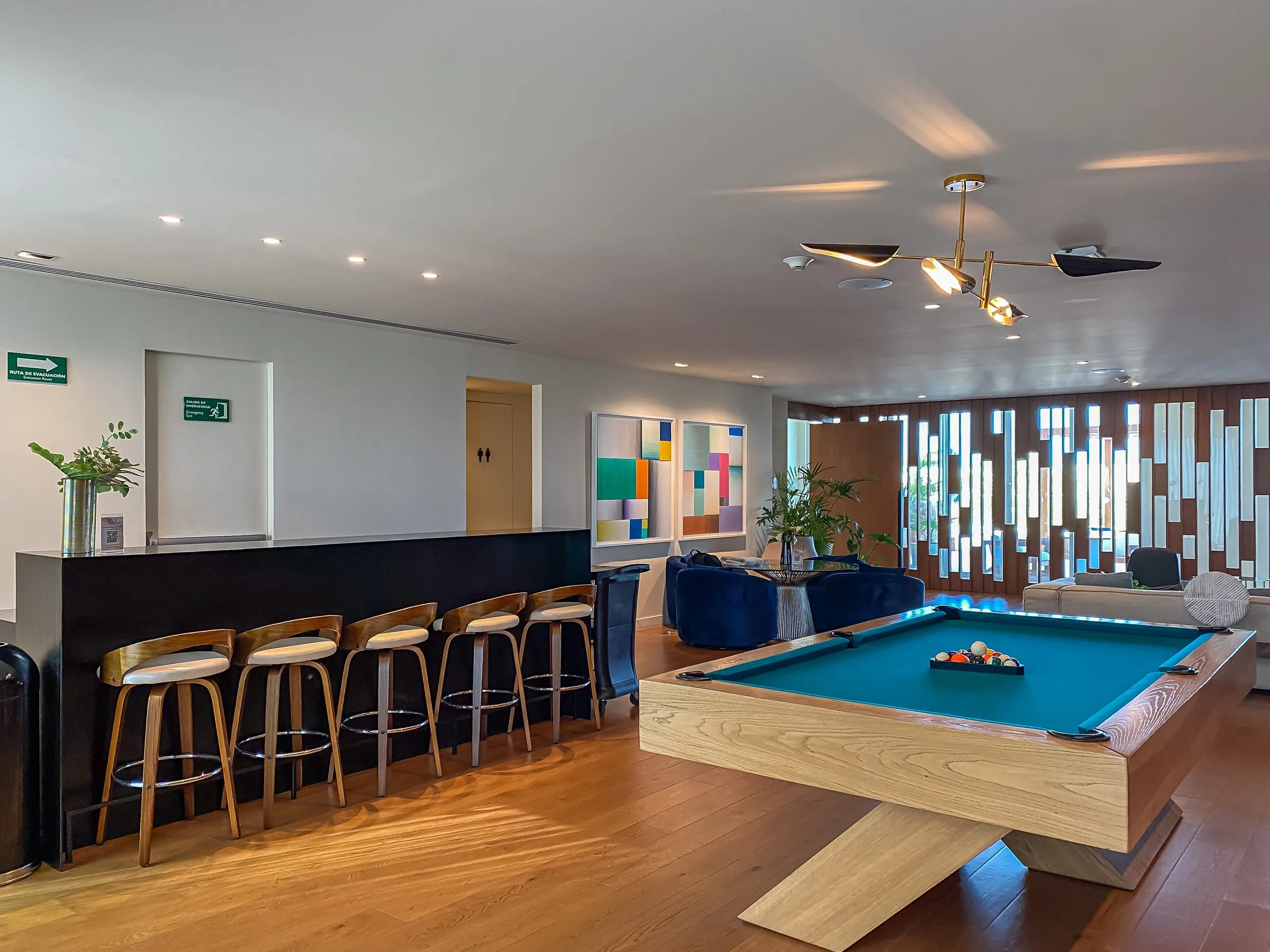 Hotel game room with a pool table, a bar with wooden stools, and blue sofas beneath a brass pendant light.