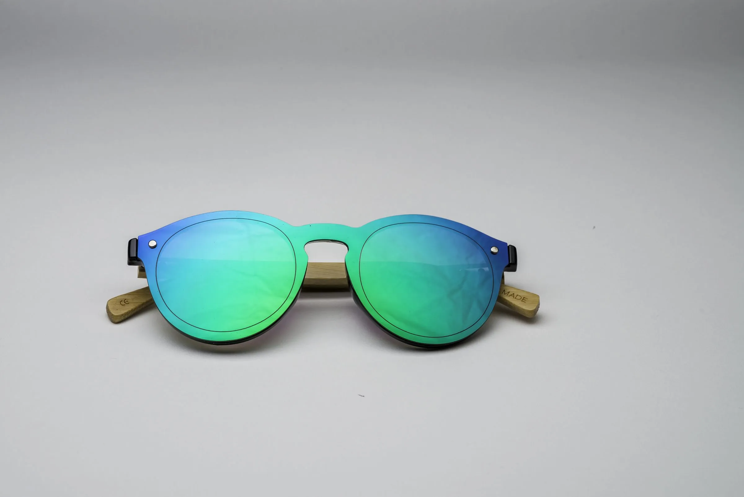 Round sunglasses folded and viewed head-on, with green-blue mirrored lenses, a black frame with a wood accent at the bridge, and wooden temple arms folded inward.