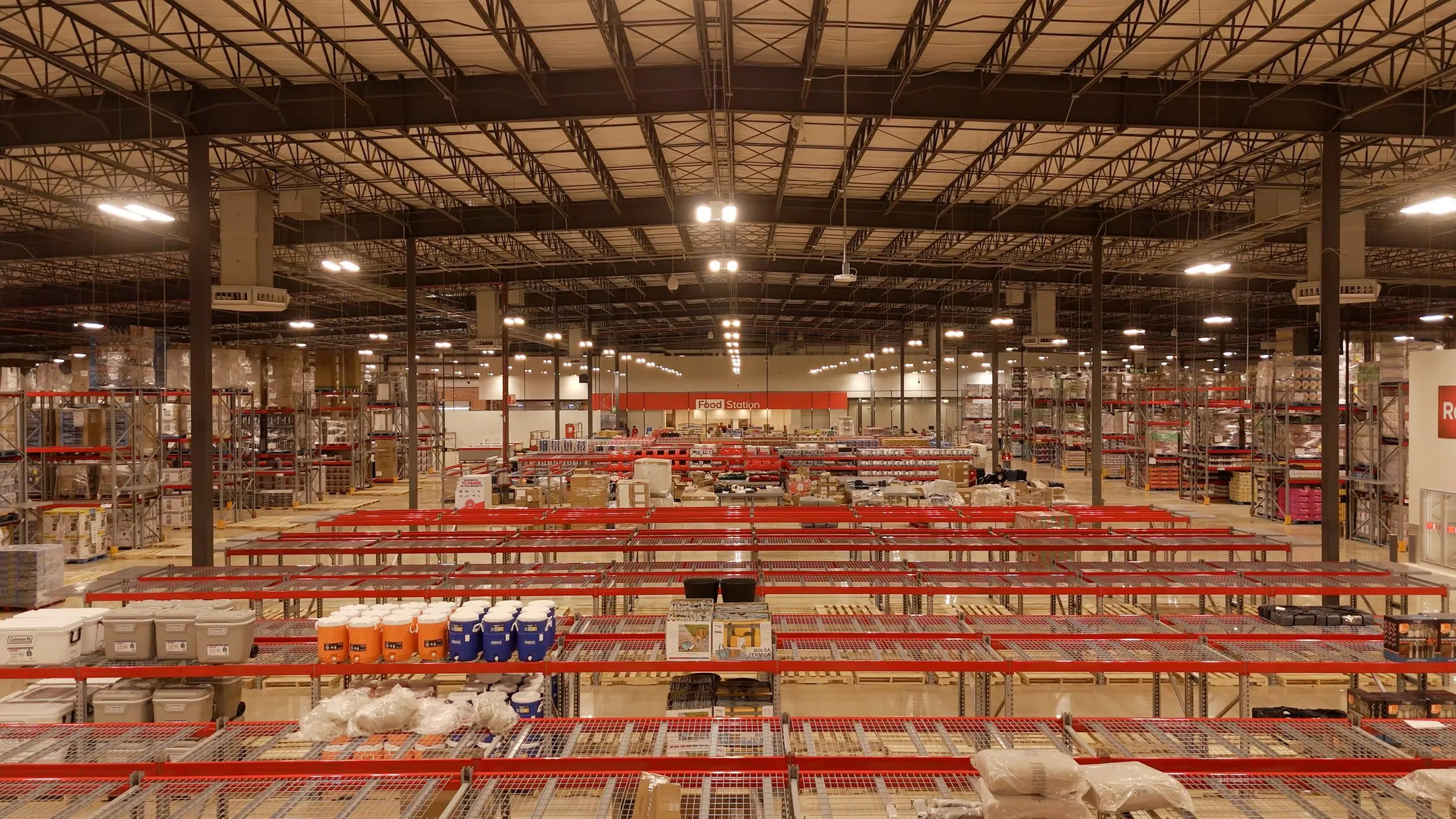 Interior of a retail warehouse with red beamed pallet racks stocked with stacked coolers and thermoses, a Food Station sign visible in the background